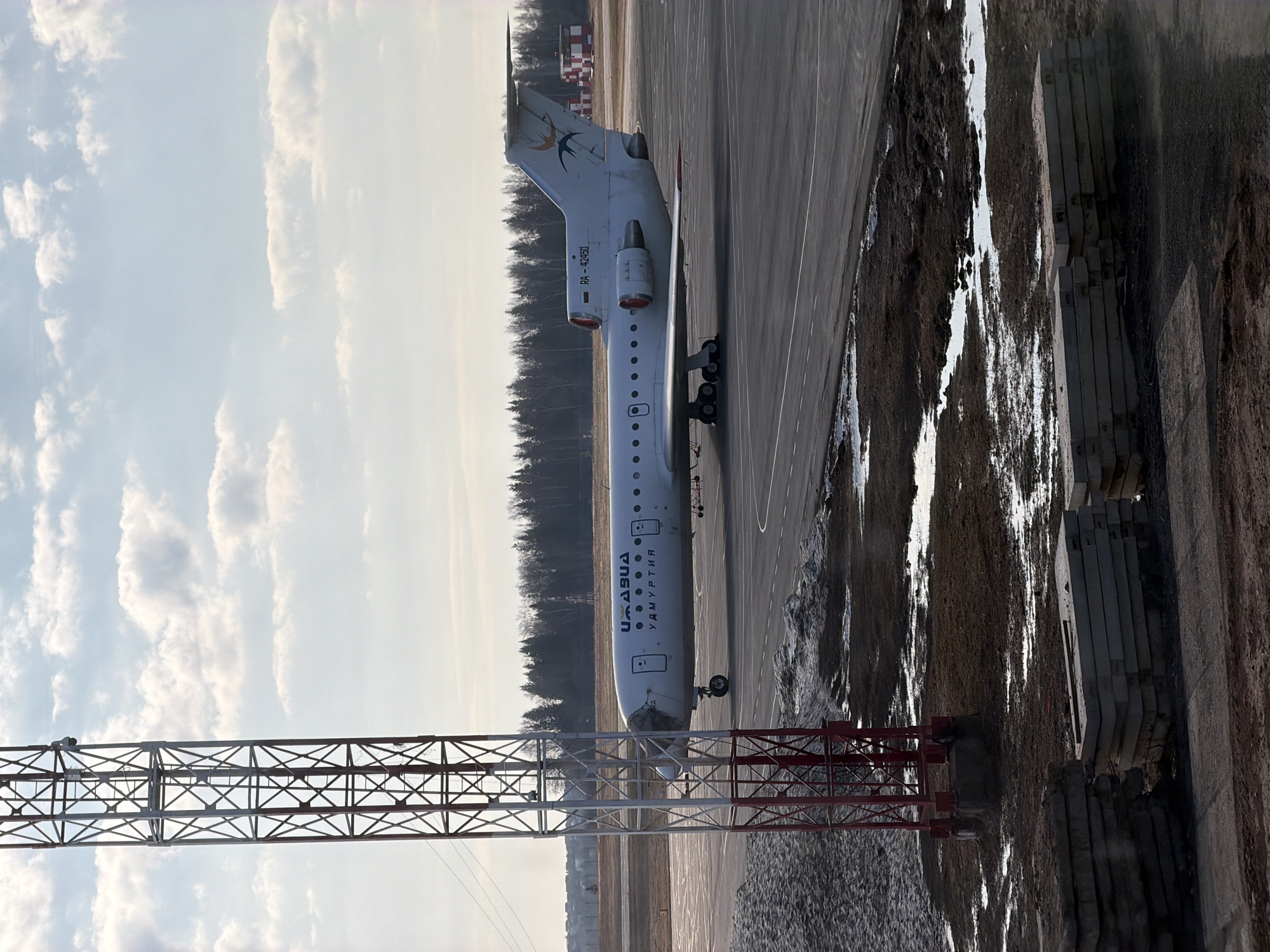 An Izhavia Airlines Tupolev Tu-134 aircraft, registered as RA-42401, is parked on an airport tarmac in Izhevsk, Russia. The white aircraft features "ИЖАВИА" (IZHAVIA) and "УДМУРТИЯ" (UDMURTIA) in dark blue lettering on its fuselage, and a stylized bird logo on its T-tail. Its twin engines are visible mounted on the rear fuselage.
The scene is set during daytime, under a bright, cloudy sky. The ground adjacent to the tarmac shows patches of snow and mud, indicating cold weather conditions, likely late winter or early spring. A dense treeline forms the distant horizon behind the aircraft. In the far background to the right, a red and white striped tower or building is visible.
A prominent metallic lattice structure, possibly a power line tower or part of airport infrastructure, stands in the foreground on the left side of the image. To the right of the tarmac, the ground is unpaved and muddy, with several rectangular concrete slabs neatly stacked. The aircraft is stationary, suggesting it is either parked or in the process of preparing for departure or arrival.
