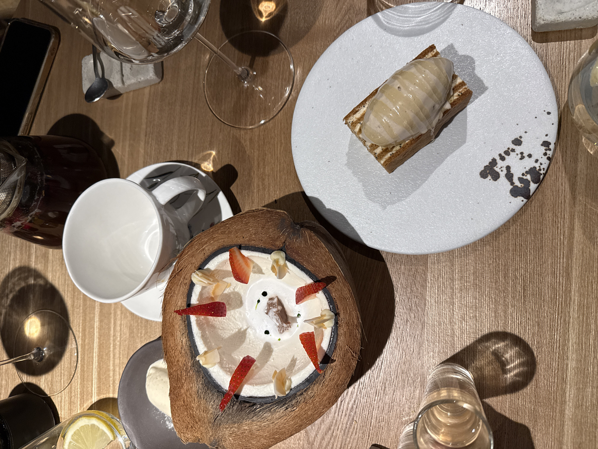 The image depicts a table setting with a variety of dishes and beverages. A coconut shell filled with a dessert, decorated with strawberry slices and white flakes, sits prominently in the center. Next to it is a white tea cup and saucer. A rectangular dessert, topped with a scoop of light-colored mousse, is placed on a white plate with dark spots. There are also glasses and a teapot on the table, along with a smartphone. The table is made of light-colored wood. The setting suggests a meal or tasting at a restaurant. There is no definitive evidence to pinpoint the location as Krasnoyarsk, Russia.