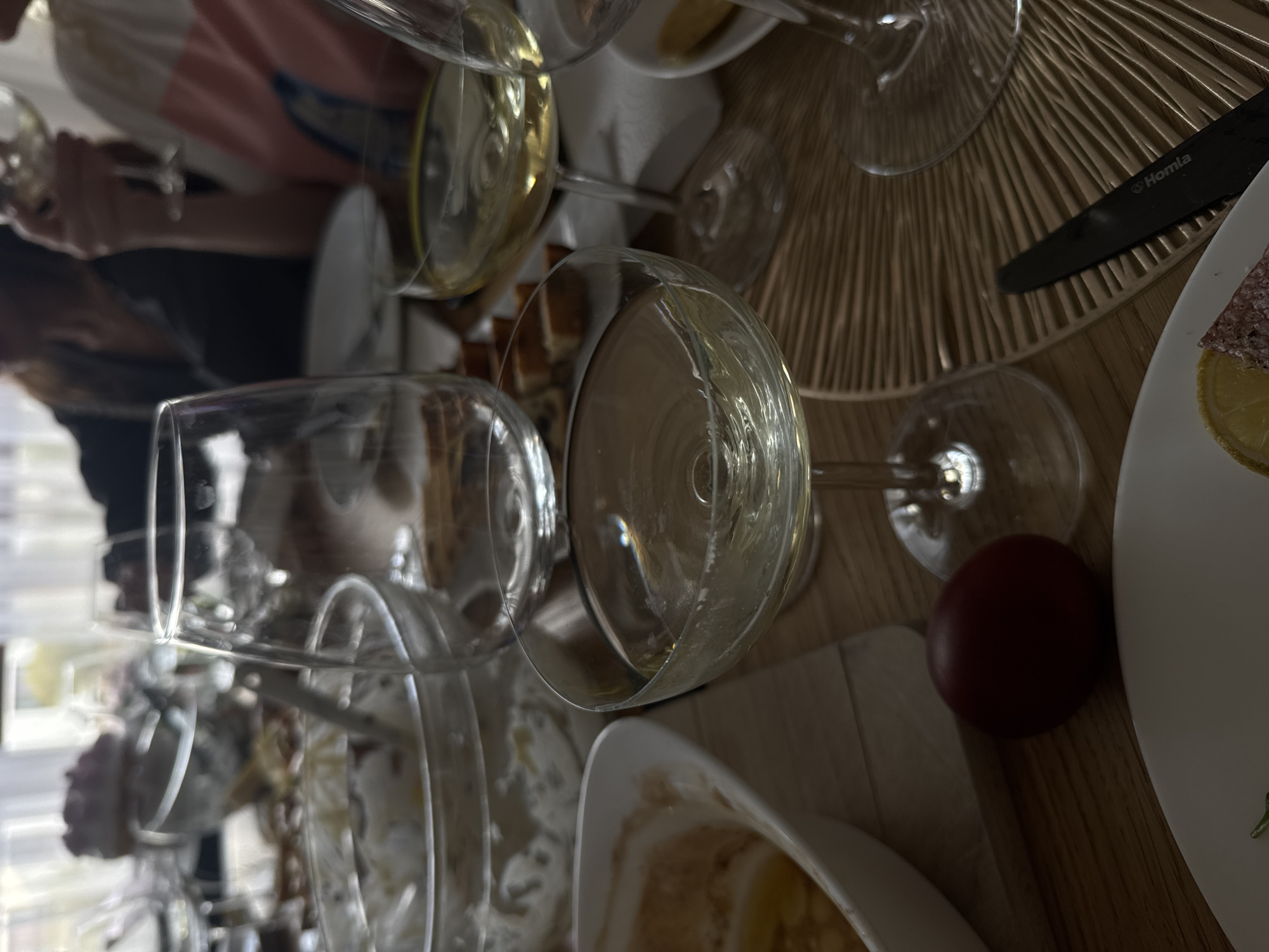 The image depicts a table setting, possibly in a restaurant or dining area, with several wine glasses, some containing liquid, possibly white wine. Plates with food are present, including what appears to be charcuterie and lemon slices. A black knife with the inscription "Homla" is visible on a woven mat. A small, dark red object, possibly a tomato or radish, is near one of the plates. Part of a person's arm and striped shirt are visible in the background. Based on the "Homla" brand, which is associated with home goods in Poland, and the general aesthetic, the image could plausibly be taken in a restaurant or dining area in a Polish city. While it is not possible to verify the specific location, the city of Warsaw, Poland is a possibility.