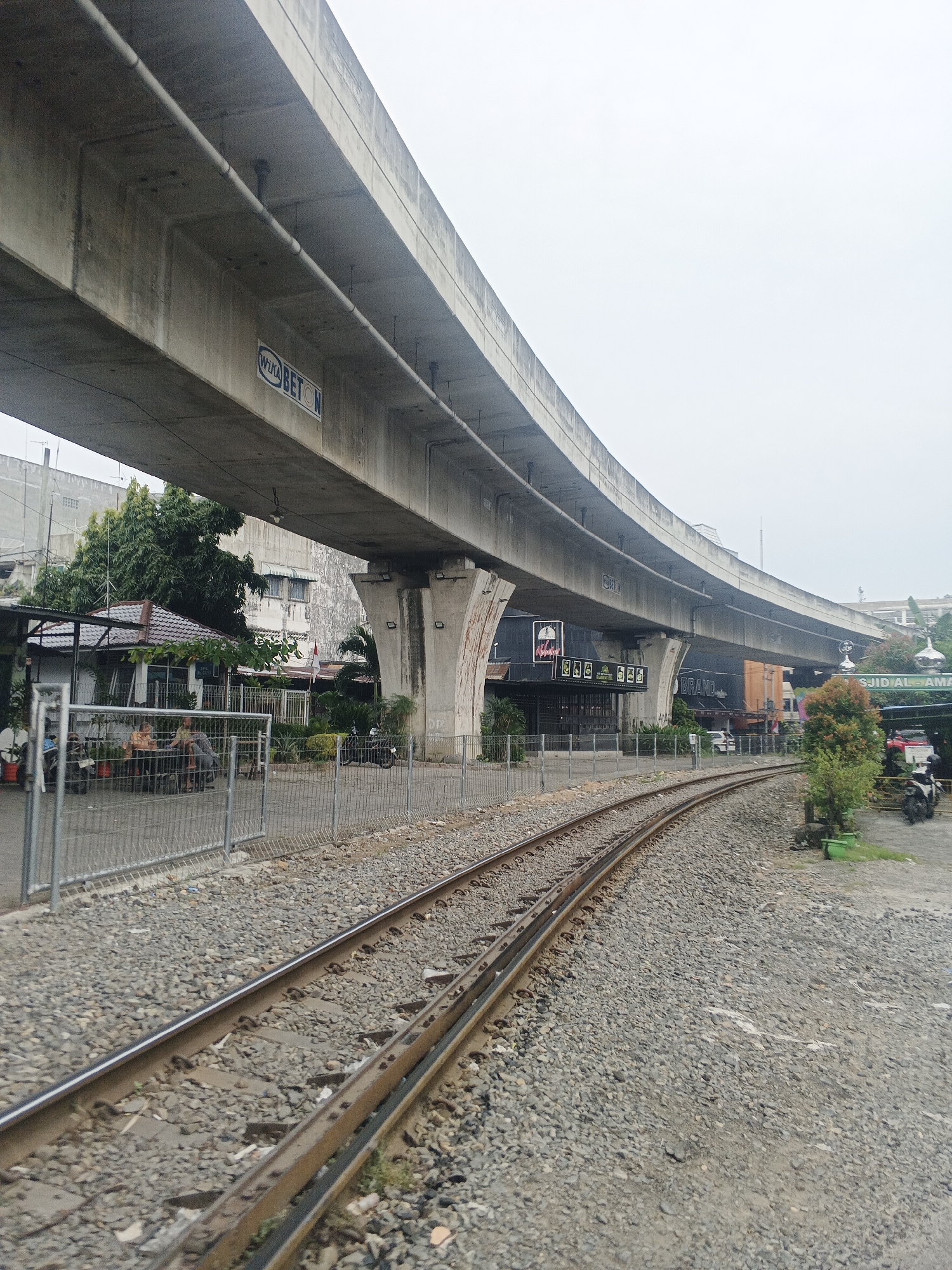 The image captures a scene in Medan, Indonesia, featuring a curved elevated concrete highway overpass supported by large pillars. Beneath the overpass, a set of train tracks curves to the right. The tracks are laid on a bed of grey gravel. Adjacent to the tracks, a metal fence encloses an area with a small structure resembling a shop or food stall with a tiled roof. Two individuals are seated at tables outside this structure. Beyond the fence and the train tracks, several buildings are visible, some appearing to be commercial establishments with signage. One sign clearly reads "SJID AL-AM." The sky is overcast. Various vehicles, including motorcycles and a car, are present in the background. Lush green trees and foliage are scattered throughout the scene.
