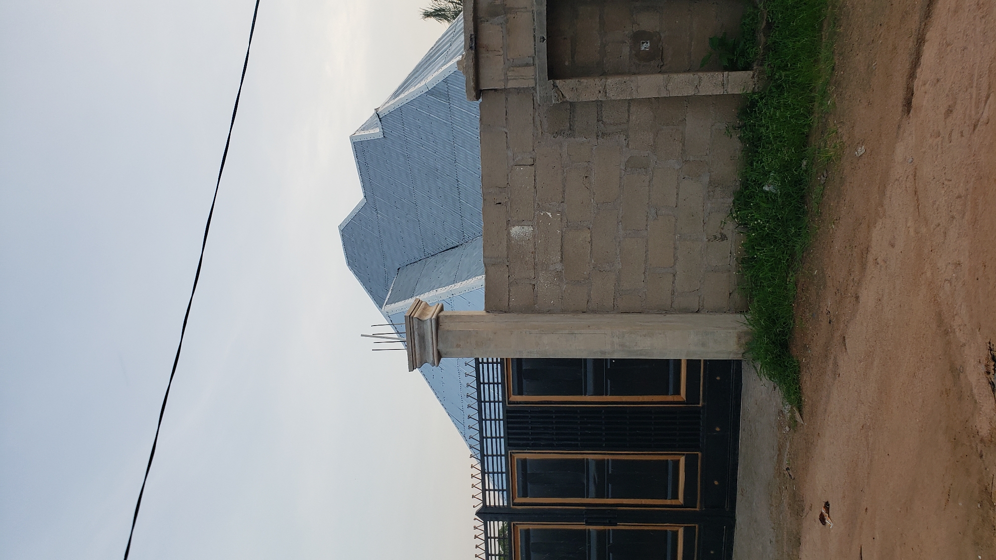 The image depicts a street view in Jalingo, Nigeria. In the foreground, a dirt road with some scattered leaves is visible. To the left, a black metal gate with gold trim stands. Behind the gate, part of a building with a corrugated metal roof is visible. To the right of the gate, a concrete pillar and a wall constructed of concrete blocks form a barrier. Green grass grows along the base of the wall. The sky above is clear and pale.