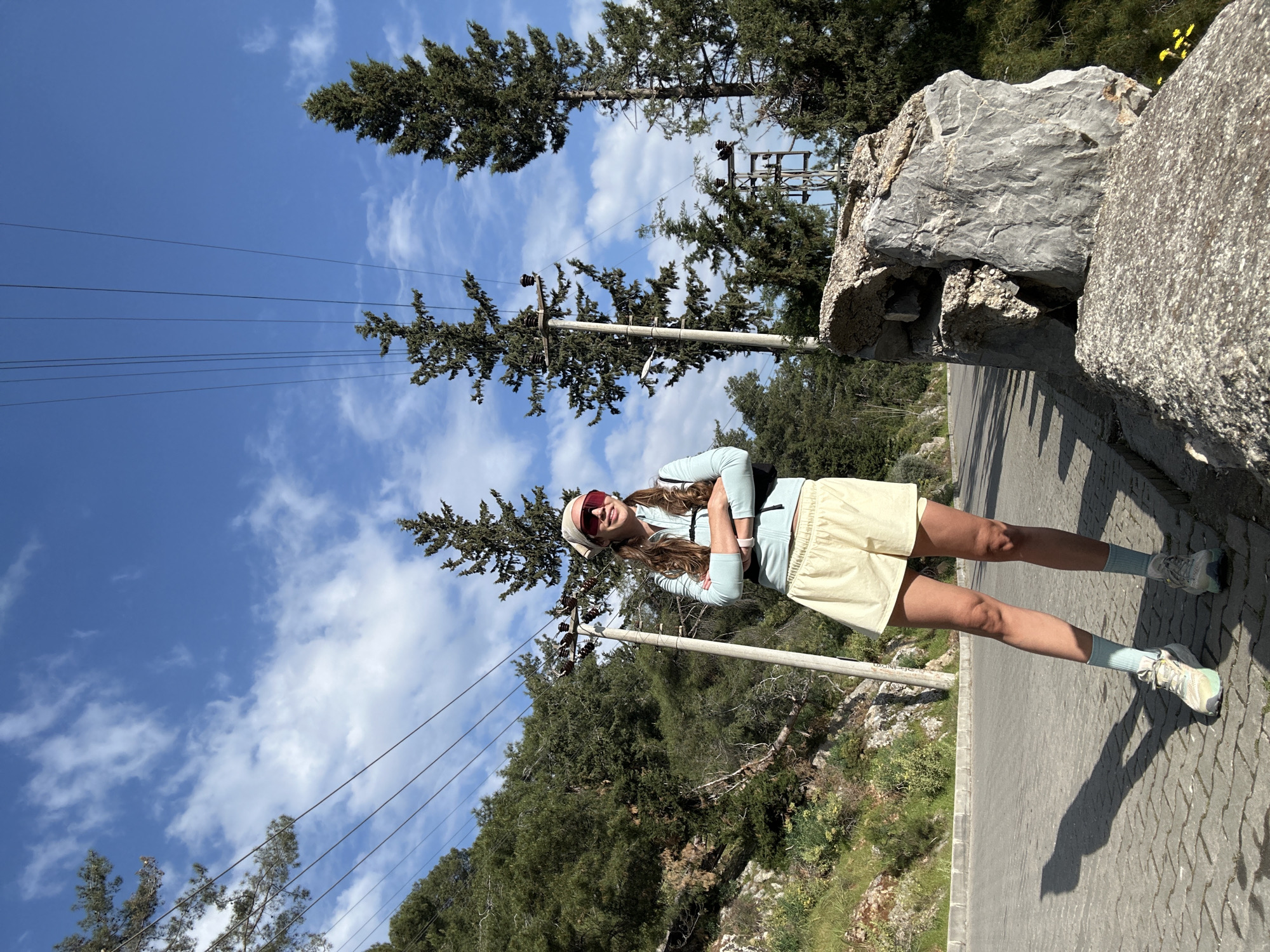 A full-length portrait of a woman is set against a backdrop of trees, a blue sky with scattered clouds, and power lines. She stands with her arms crossed, wearing a light blue top, pale yellow shorts, red-tinted sunglasses, and a headband. She also has on light blue socks and running shoes. To her right, part of a stone structure or wall is visible along a paved road. The setting appears to be outdoors in a mountainous area, likely in the vicinity of Fethiye, Turkey.