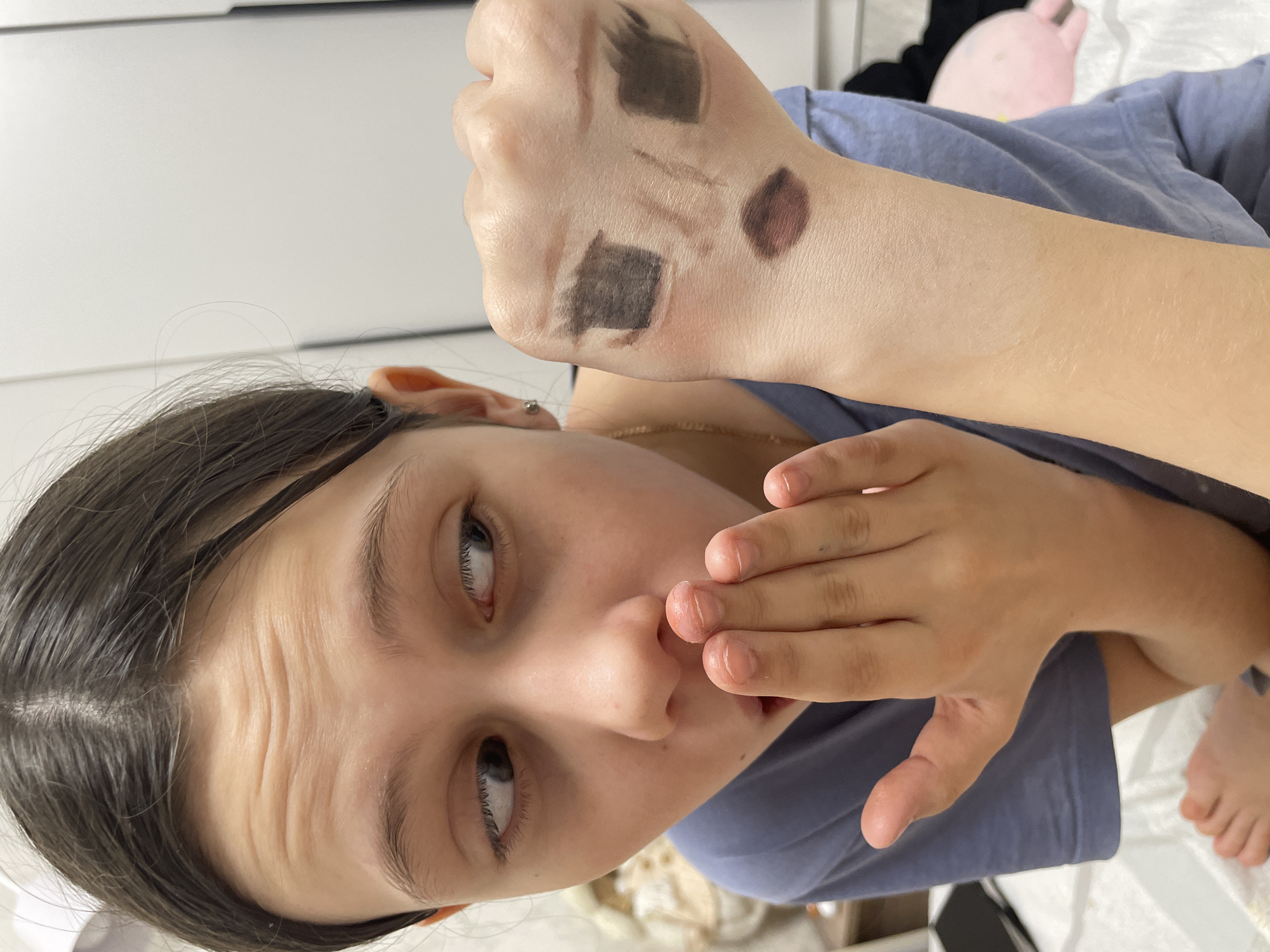 The image captures a close-up, diagonal view of a young person, likely female, engaged in testing makeup products. The subject is looking to her right with an expression that appears curious or slightly surprised, with her eyebrows subtly raised and her mouth partially covered by her left hand.
Her dark hair is pulled back, revealing her forehead which shows natural creases. She has light-colored eyes, possibly blue or grey, which are directed upwards and to the right. A small silver stud earring is visible in her left earlobe, and a thin gold chain necklace rests around her neck. She is wearing a simple light blue or lavender t-shirt.
The primary action depicted is the testing of makeup. Her right hand is held up, showcasing several swatches on the back of it. These swatches consist of various shades of brown, taupe, and dark grey, suggesting they are eyebrow products, eyeshadows, or contour shades. There are at least five distinct swatches: two larger, squarish patches, one diamond-shaped swatch, and two thinner, linear strokes.
The background is minimalist, consisting mainly of a bright, light-colored surface, possibly a wall or a piece of furniture. In the top right corner of the image, hints of a pink object, possibly a blanket or soft toy, and a bare foot are visible. The lighting is bright and even, indicating an indoor setting. The image conveys a personal, everyday moment of experimenting with beauty products.