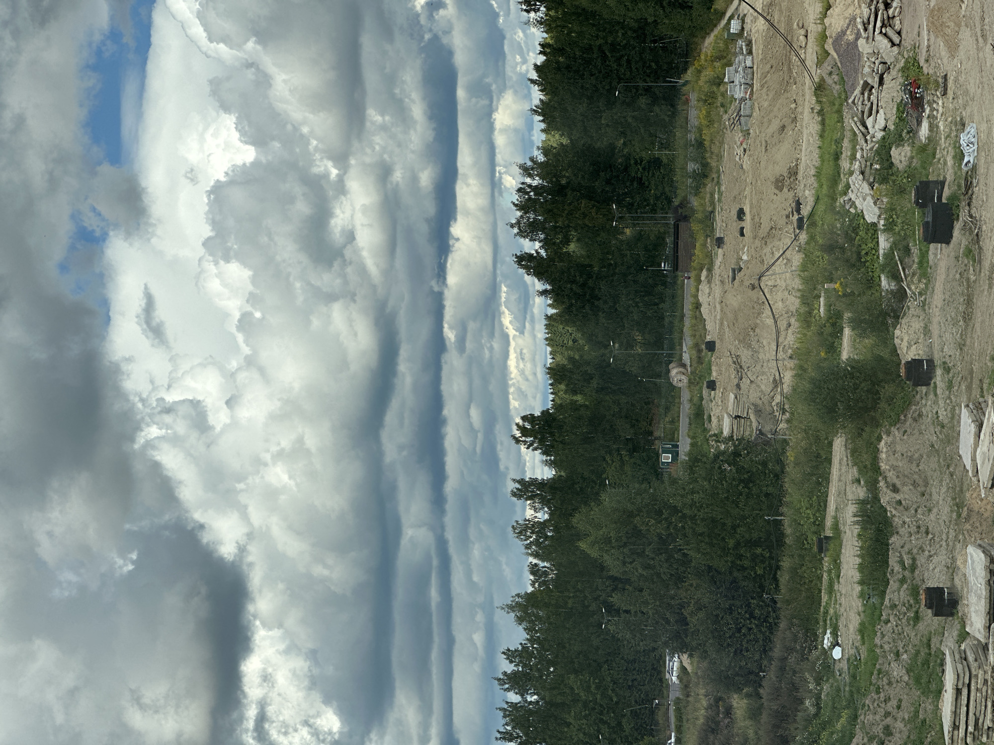 The image depicts an outdoor construction site with a dense forest in the background, under a dramatic sky filled with dark, layered clouds. In the foreground, the construction site shows mounds of dirt, piles of concrete slabs, and black cylindrical objects that appear to be part of a foundation. Various cables and construction materials are scattered across the sandy terrain. A road is visible on the left side of the image, with two white cars parked near the edge of the forest. Several lampposts are also present within the construction area and along the road. A small green utility box and a large wooden spool are visible further into the site. The scene suggests ongoing construction activity in a semi-rural or suburban area, possibly on the outskirts of Vsevolozhsk, Russia, given the presence of a characteristic Russian forest.