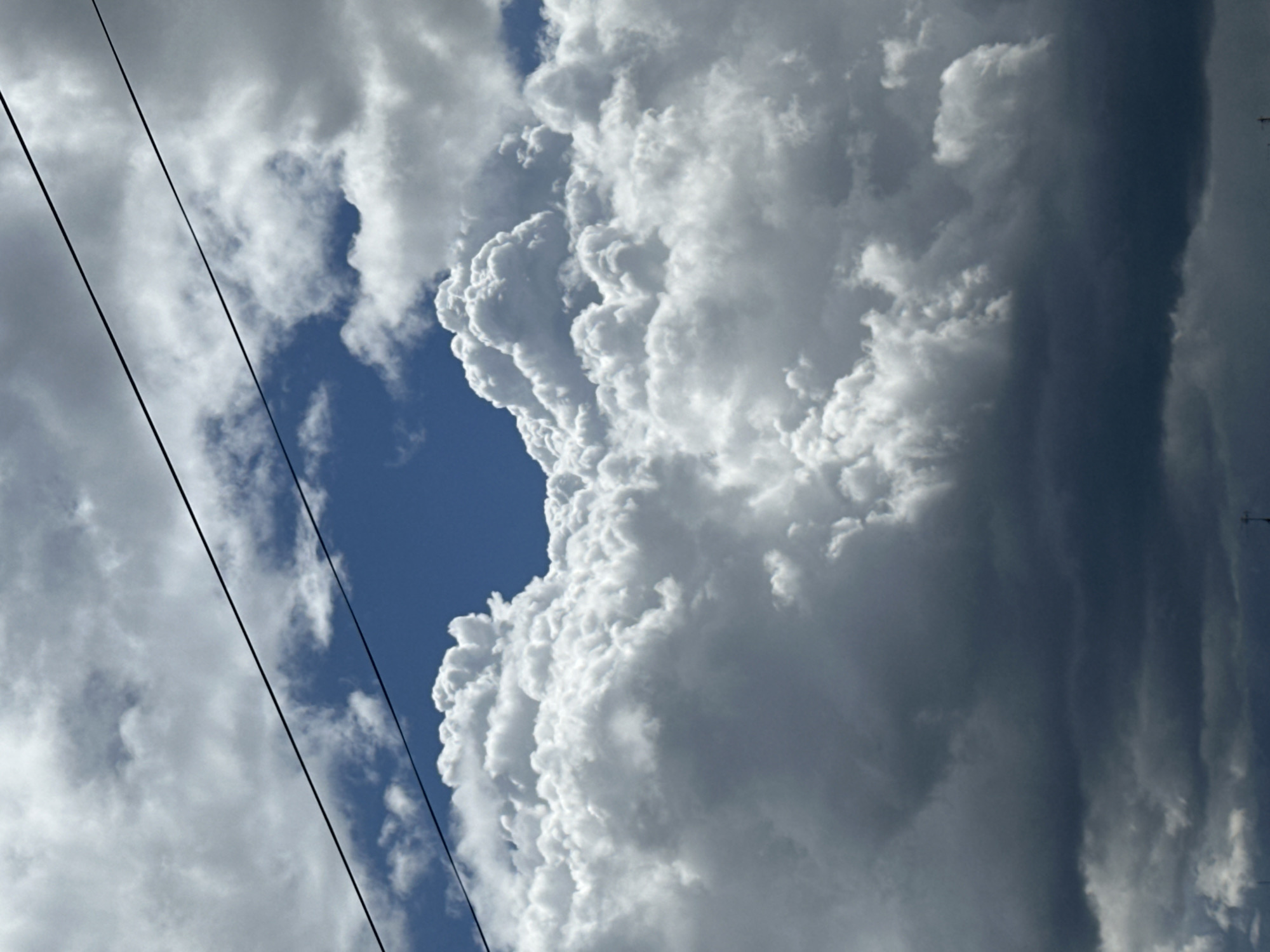The image shows a view of the sky, presumably in an urban area given the presence of power lines. The sky is mostly cloudy, with a patch of clear blue visible amidst the clouds. The clouds are dense and appear to be cumulonimbus or similar cloud types, with a bright, billowing texture. To the right, darker, rain-bearing clouds suggest the possibility of an approaching storm. The location is Unknown, Unknown. There are no people in the frame. The primary objects are clouds and power lines. The environment is atmospheric, with a focus on weather conditions. There are no specific actions or interactions depicted.