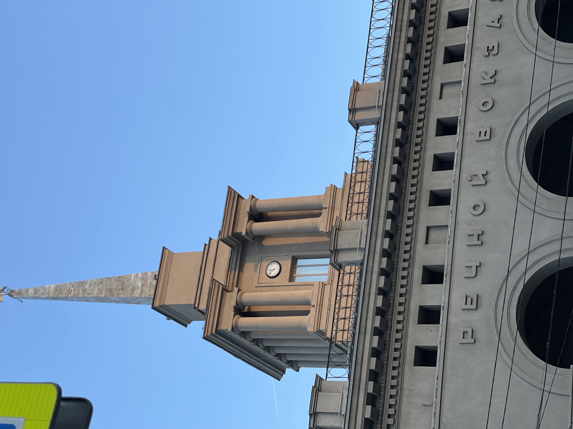 The image displays a grand, monumental building, characteristic of Stalinist Empire architecture, captured from a low angle looking upwards against a clear, bright blue sky. The structure is built from light grey or beige stone/concrete, featuring elaborate classical elements such as numerous columns, decorative cornices, and recessed rectangular window openings.
A prominent, slender spire extends from the building's highest point, tapering to a sharp tip, possibly adorned with a small golden star. Below the spire, an ornate section of the tower includes a circular clock face positioned above a window, framed by sets of robust columns. Intricate metal railings with circular motifs line the edges of different levels of the building.
On the main facade, large, raised Cyrillic letters spell out "ВОКЗАЛ" (VOKZAL), which translates to "station" in Russian, clearly indicating its function, likely as a railway or bus station. Multiple dark cables run vertically over this part of the facade, intersecting large circular architectural features.
The sky is a vibrant, cloudless blue, suggesting a sunny daytime scene. A faint airplane contrail is visible in the distance. In the bottom-left corner, a partial view of a yellow and black striped object, possibly part of a traffic or pedestrian crossing sign, is present. The building is located in Krasnoyarsk, Russia.