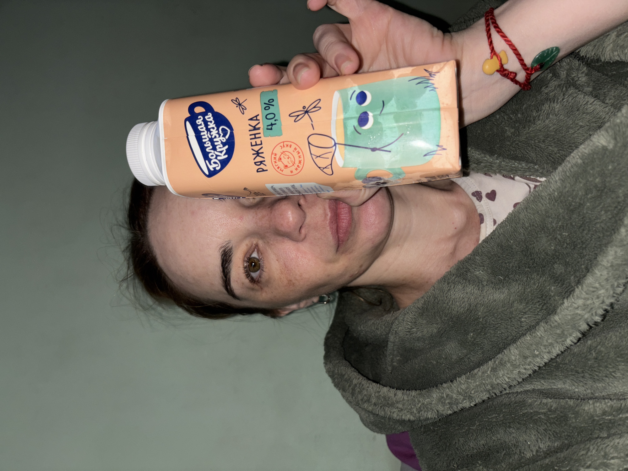A woman indoors, with her face visible, holds a carton of "Ryazhenka" drink on her forehead. The carton is peach-colored with text in Russian and a cartoon illustration of a cup with a face on it. The woman is wearing a gray fleece jacket. A red bracelet with charms is visible on her wrist. The background is a plain wall. The location is Unknown, Russia, inferred from the Russian text on the carton.