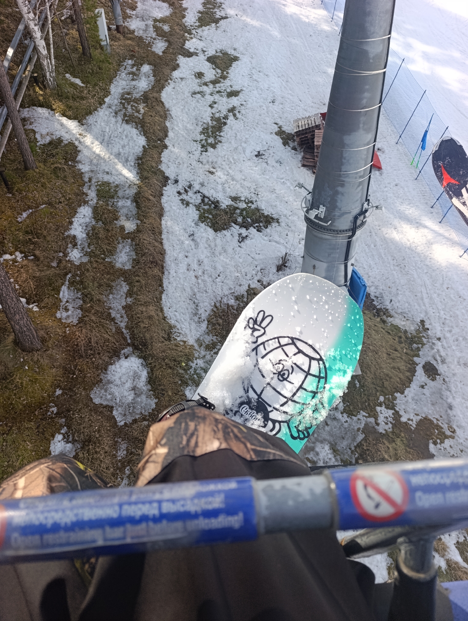 The image shows a first-person perspective from a ski lift, likely at a ski resort in Krasnoyarsk, Russia, based on the Cyrillic lettering on the safety bar in the foreground which translates to "Open restraining bar just before unloading!". The view is downwards at a snowboard resting on the chairlift. The snowboard has a white base with an illustrated cartoon globe design and the brand "Cukorka". The rider's legs, clad in camouflage pants, and boots are visible below the safety bar.
The background reveals a partially snow-covered landscape with patches of grass and dirt. A ski lift support tower is positioned to the right of the snowboard. In the distance, a groomed ski run can be seen. Fencing and what appears to be a red and black snowboard are also visible on the run. The weather appears to be clear and sunny.