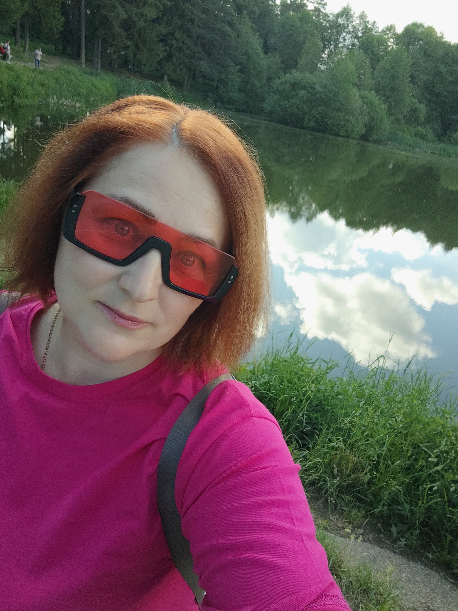 A woman is taking a selfie outdoors near a body of water. She has reddish-orange hair and is wearing large, black-framed sunglasses with red lenses and a bright pink t-shirt. A shoulder strap of a bag is visible on her shoulder. The water reflects the sky, including clouds. There is green grass and trees in the background. Based on the image, it's likely the location is in a park or natural area in the city of Chaikovsky, Russia, although this cannot be confirmed.
