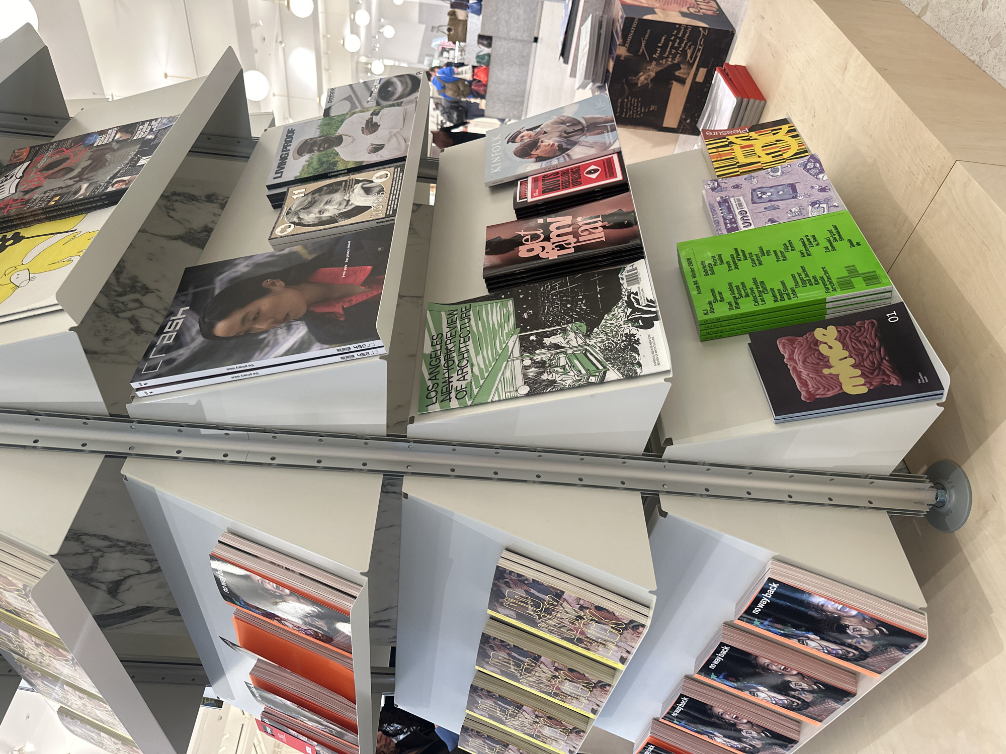 The image shows a display of magazines on angled shelves. The shelves are made of white metal and have a marble-like finish on the inside. The magazines cover a variety of topics, including architecture, fashion, and culture. Titles include "Crash", "Living Proof", "Kinfolk", "Los Angeles, New York Review of Architecture", "Pleasure", and "No Way Back". The display is located in a store in London, United Kingdom. Several people can be seen walking past the shelves in the background. The lighting is bright and even.