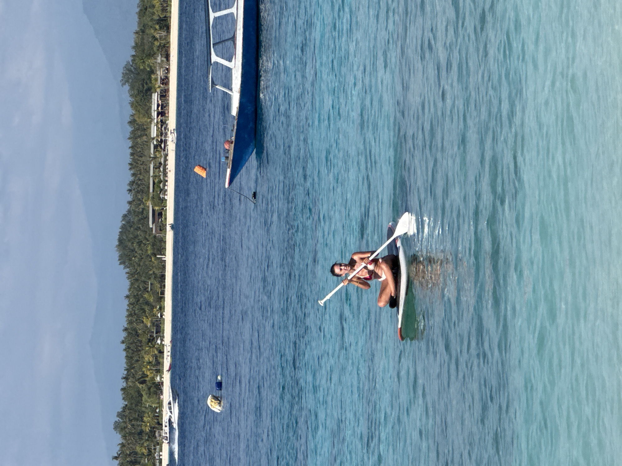 A woman is paddleboarding on the ocean near an island in Indonesia. She is wearing a red and white bikini and sitting cross-legged on the paddleboard. She is holding a paddle. There are two boats in the water. One is blue and white and closer to the shore. The other is white and further out to sea. In the background, there is a white sandy beach lined with palm trees. Mountains are visible in the distance.