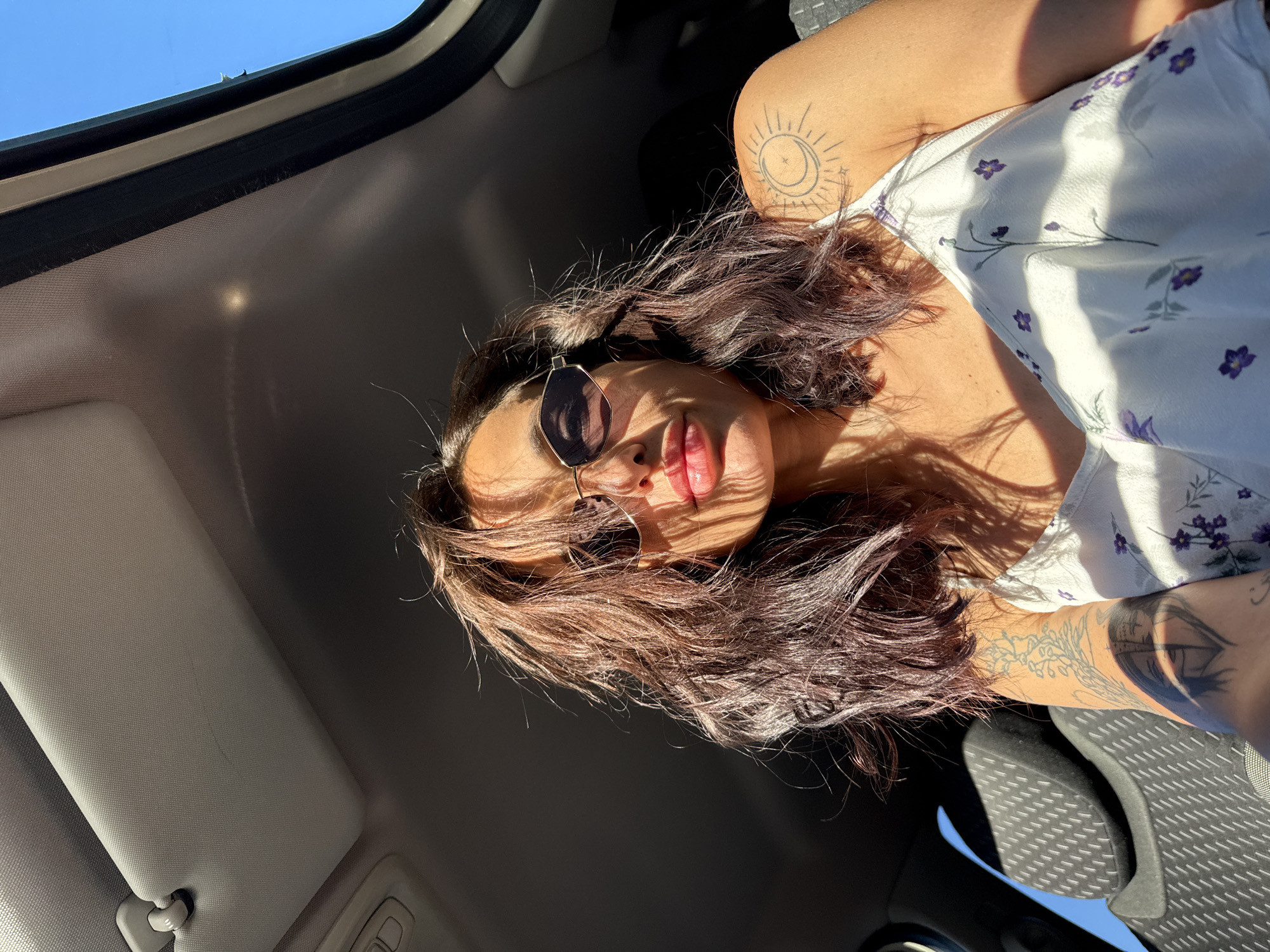 A woman is photographed from a low angle inside a car, looking up at the camera. She has dark, wavy hair and is wearing sunglasses. Sunlight streams in from the right, casting shadows across her face and the interior of the car. She is wearing a white dress with a floral pattern. Tattoos are visible on her arm and shoulder. The interior of the car includes a sun visor and the back of a seat. The scene appears to be a sunny day, possibly in the city of Pleven, Bulgaria, given the context provided.