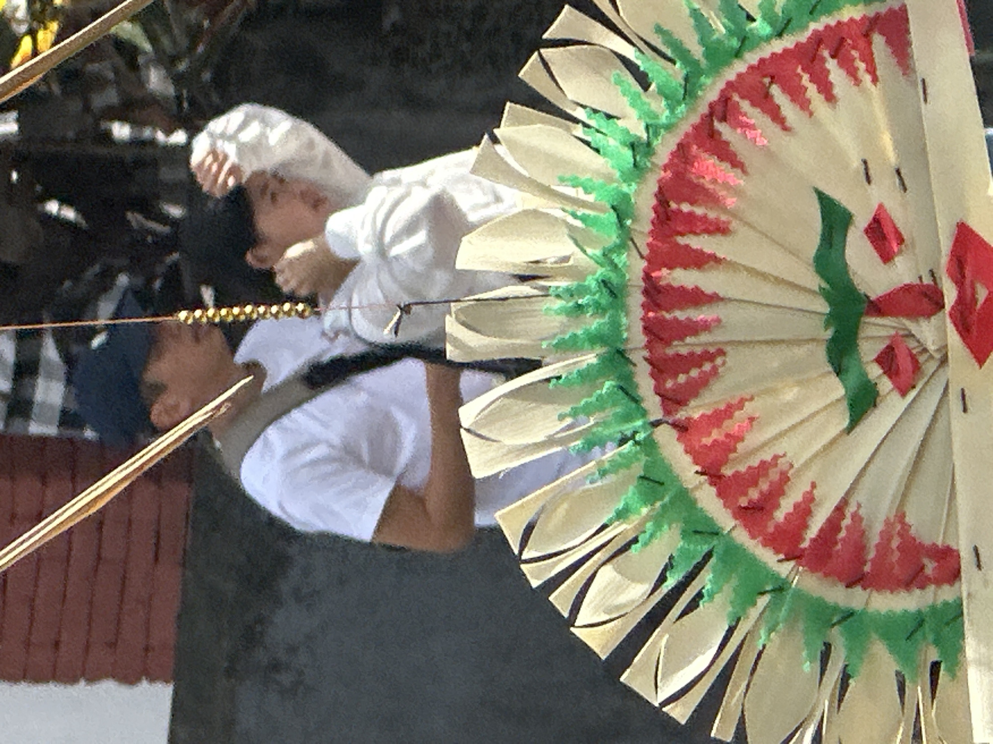 The image shows a close-up view of a person from the chest up, wearing a white t-shirt and a dark baseball cap. Behind them, another person is partially visible, also wearing white clothing. They appear to be standing outdoors, with a blurry background of foliage and a brick wall. In the foreground, a large, decorative fan made of palm leaves is prominently displayed. The fan is intricately folded and painted with red and green patterns, resembling a traditional Balinese headdress or offering. The scene is brightly lit, suggesting it is daytime. The overall impression is of a cultural event or ceremony taking place in Ubud, Bali, Indonesia.