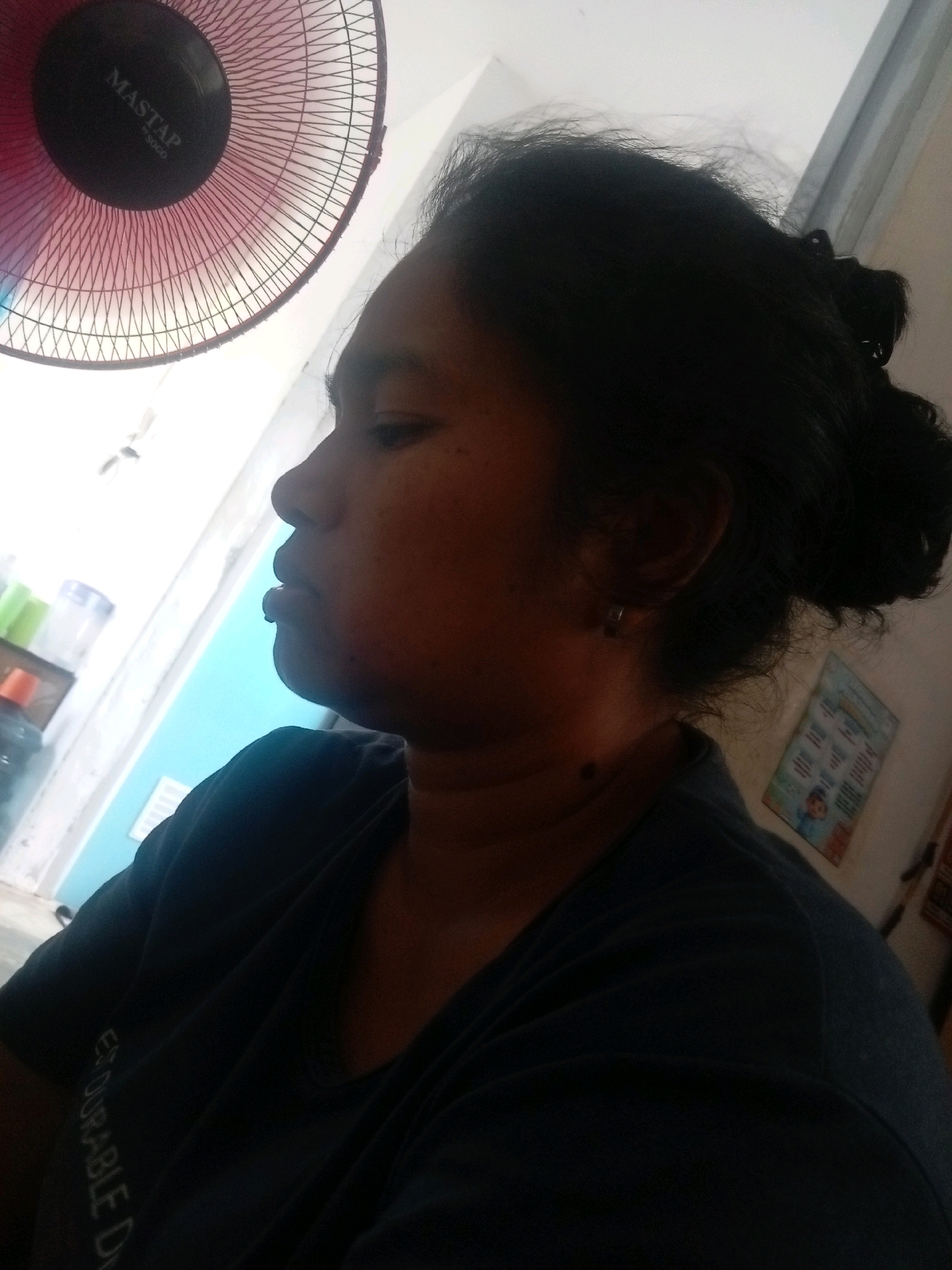 The image shows a close-up profile view of a woman with dark hair pulled back in a bun. She is wearing a dark t-shirt with text on the left side that reads "EXODURABLE D". A portion of a red oscillating fan with the brand "MASTAP" is visible in the upper left corner. The background suggests an indoor setting with a light-colored wall and a bright window. There are some indistinct objects on a shelf in the left background, including what appears to be a water bottle. A blue door or wall is also visible in the background. The overall lighting is bright. The image was taken in Batam, Indonesia.