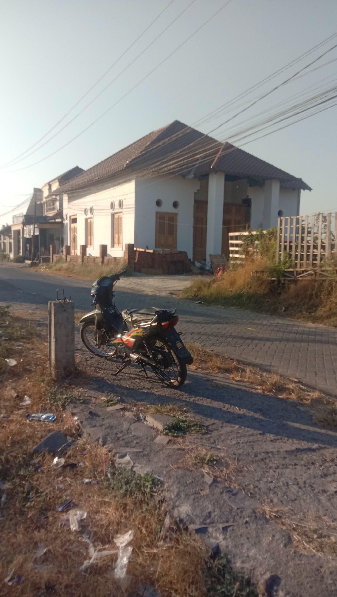 The image depicts a scene on a street in Curah Dukuh, Indonesia, under a clear, pale sky. A black motorcycle with colorful decals is parked on the side of a paved road. The road surface appears to be made of interlocking bricks. Adjacent to the road is a concrete post. In the background, a white building with a brown tiled roof is visible. The building has several windows and doors, some of which are open. The area is sparsely vegetated with dry grass and some scattered litter. Power lines are visible crisscrossing the sky above the building. The overall impression is of a quiet, possibly rural or semi-urban street.