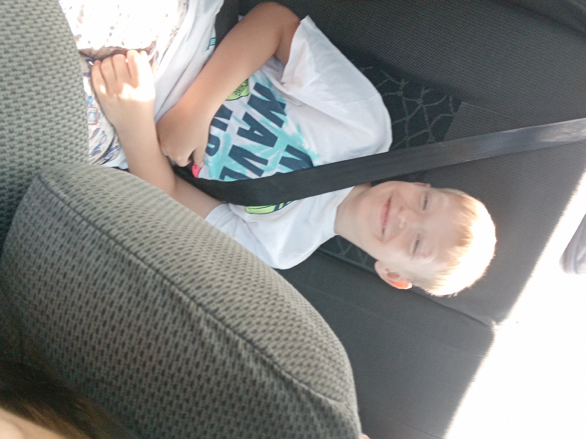 In a sun-drenched car interior, a young boy with blond hair and a wide smile is seated in the back seat, wearing a white t-shirt with a "Wave" graphic. He is secured by a black seatbelt. The seat he is in appears to be a dark gray fabric. Another person's shoulder and head are partially visible in the foreground, also seated in the car, creating a slightly blurred effect. The overall scene suggests a casual outing or road trip within the city of Kam'yana Yaruha, Ukraine, with the bright sunlight indicating it is daytime.