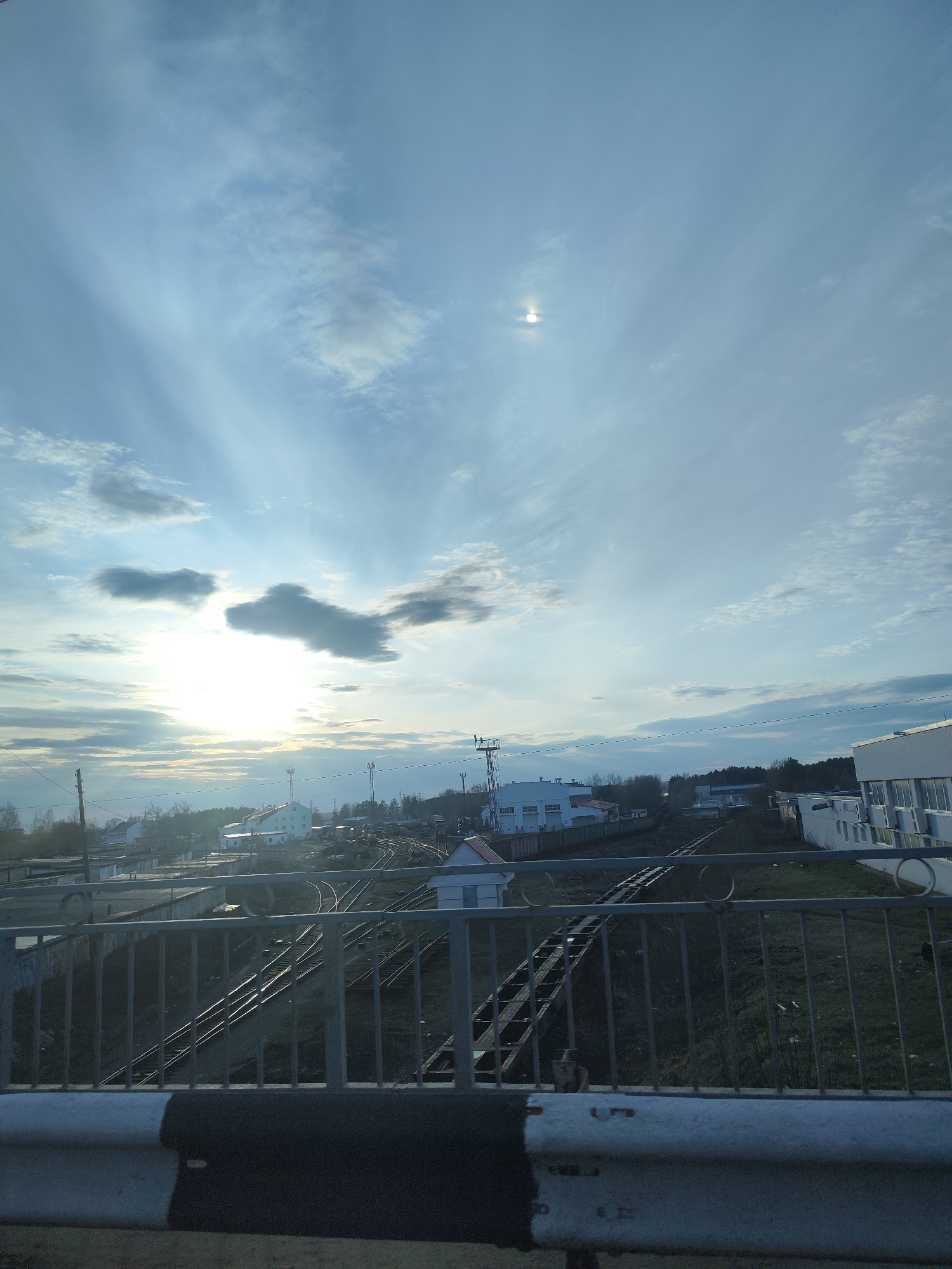 The image is a low-angle, daytime shot taken from a bridge overlooking a railway junction in Chaikovsky, Russia. The sky is mostly filled with textured clouds, with the sun visible through a break in the cloud cover on the left side of the image. A second bright light source in the sky is present high on the right side. The railway tracks extend from the foreground into the middle distance, curving and branching out. The ground between the tracks is mostly covered in dark vegetation and detritus.
Beyond the railway, several buildings are visible. A single white building with a red roof is in the mid-ground, left-of-center. Further back, a larger white industrial building is visible on the right side of the frame. The environment is industrial, with an unkempt and somewhat neglected appearance.
The bridge from which the image is taken is marked by a gray metal railing. The lower part of the frame shows the concrete barrier of the bridge, which has a black and white marking.