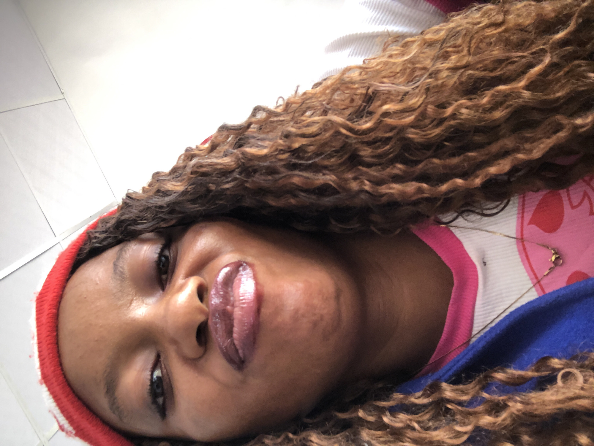 A close-up, tilted portrait of a dark-skinned woman with curly brown hair. She is wearing a red and white striped beanie. Her lips are glossy. She is wearing a pink and white top with a blue jacket. A thin gold necklace is visible. The background consists of white walls and a striped surface. The location is unknown; there is no information within the image to indicate it is in Pindiga, Nigeria.