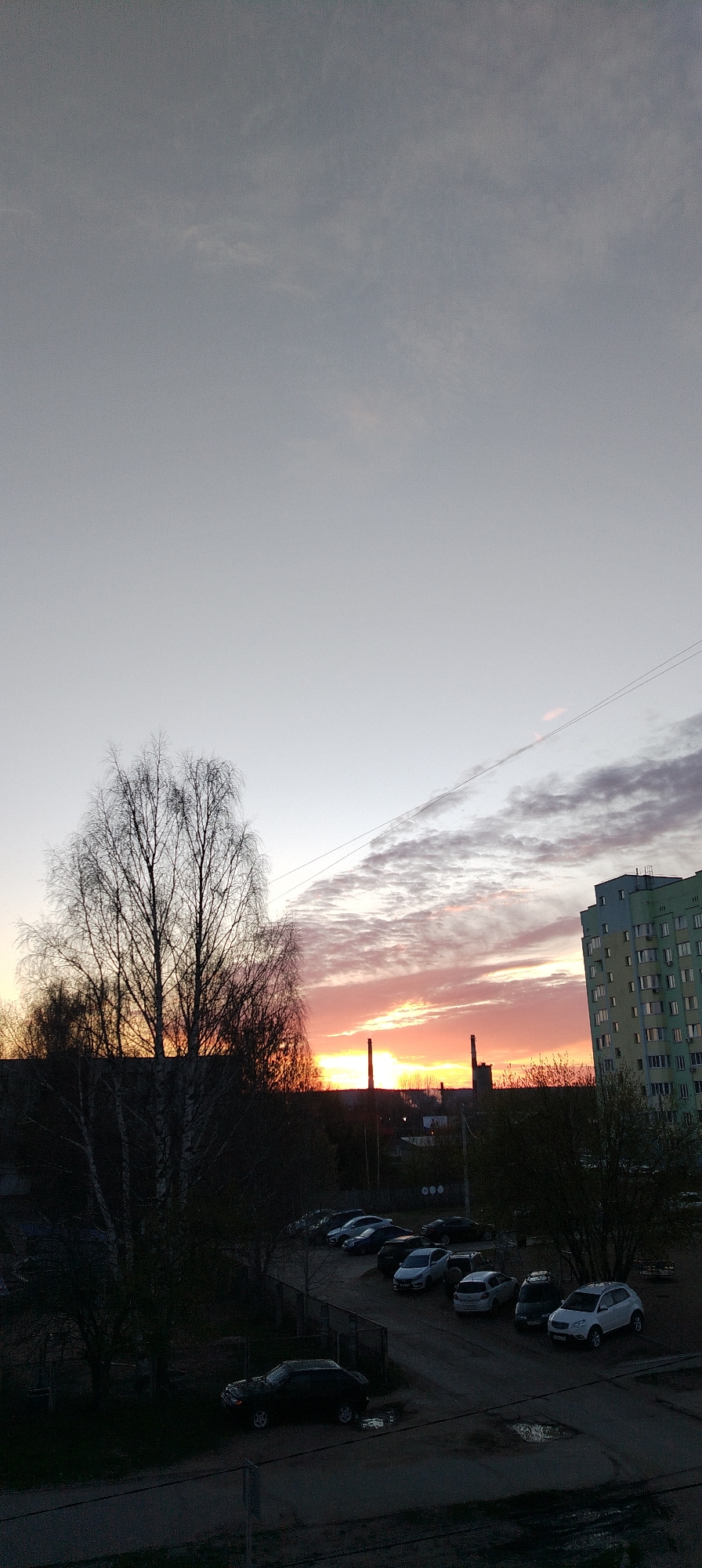 The image captures a sunset scene in an urban area. The sky transitions from a light gray at the top to vibrant orange and pink hues near the horizon. Silhouetted against the sunset are two tall industrial chimneys or smokestacks. A large building with multiple windows is visible on the right side of the frame. In the foreground, bare trees and foliage frame a parking lot containing several cars. The overall scene suggests a late evening setting in a city.
