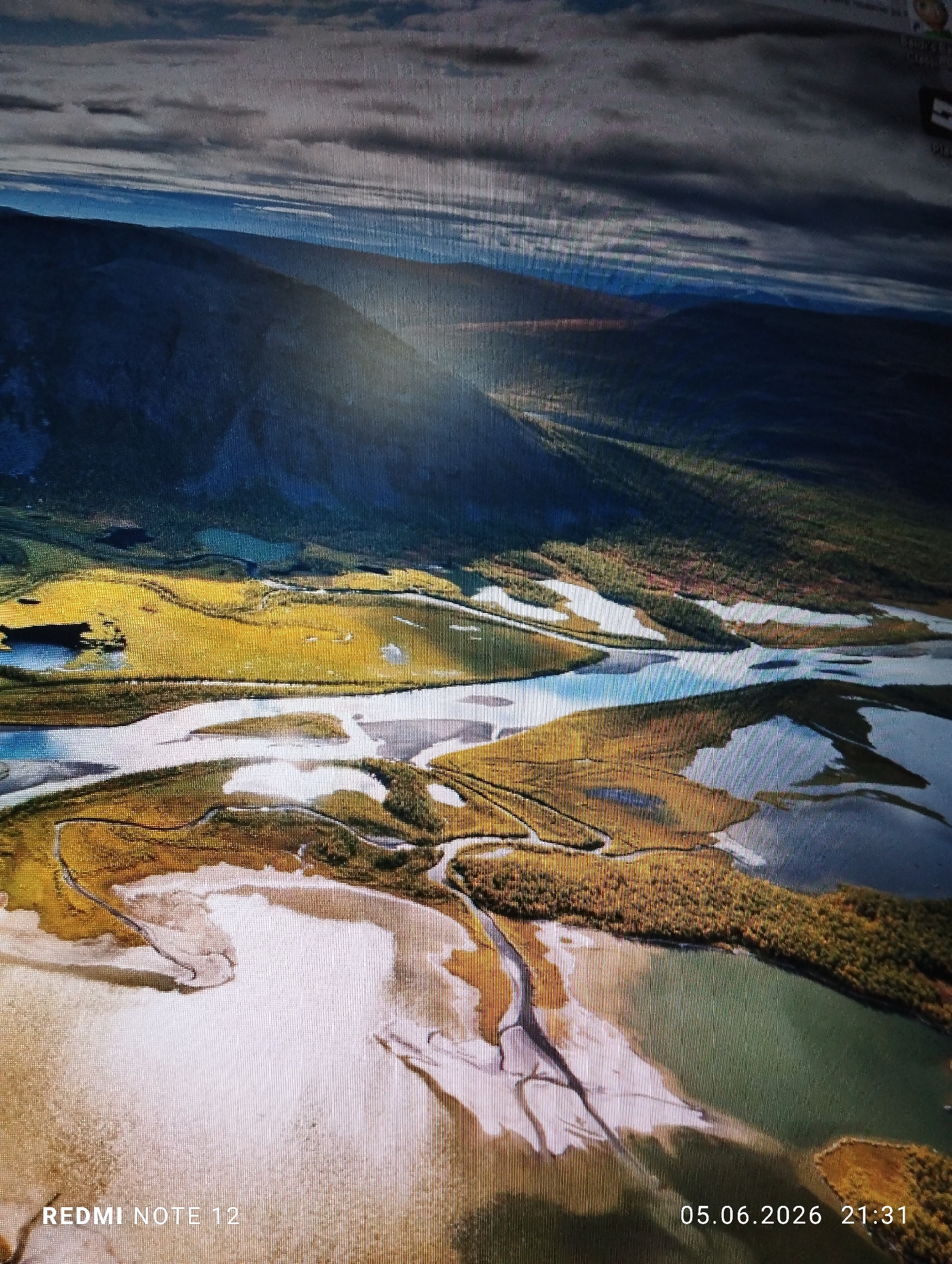 The image is a photograph capturing a digital screen displaying an aerial perspective of a natural landscape. The scene, specified as being in the city of Burgas, Bulgaria, features a meandering river system with numerous sandbanks and distinct water channels. In the upper portion, large, dark-hued mountains or hills rise, with a prominent ray of sunlight descending from the upper left, illuminating a section of the mountainside and casting light across the valley. The terrain transitions from dark green, possibly forested areas on the mountains, to extensive sections of vibrant yellow or golden vegetation, likely fields or autumn foliage, along the riverbanks. The river itself shows varying depths and sediment patterns, particularly in the foreground where broad, lighter areas suggest shallow waters or exposed riverbeds. No people, animals, or artificial structures are discernible within the landscape. The visual artifacts of the photograph, including visible vertical pixel lines, minor glare, and slight color distortion, confirm it is a picture of a monitor. Text overlays from the capturing device are present: "REDMI NOTE 12" in white in the bottom left corner and "05.06.2026 21:31" in white in the bottom right corner, indicating the phone model and the date/time the photograph was taken.