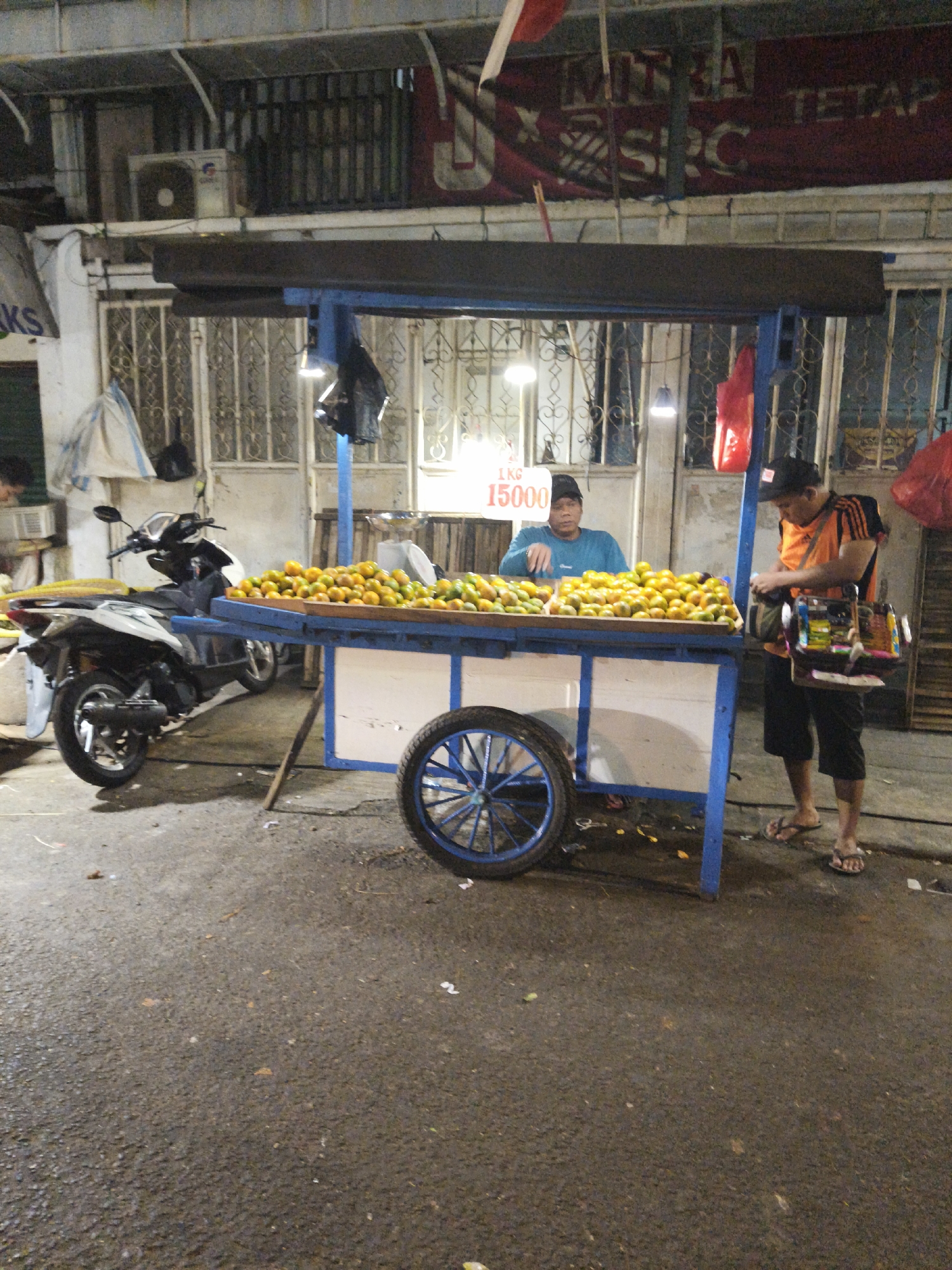 A side-view, medium shot captures an outdoor scene at dusk in South Jakarta, Indonesia, near a street vendor's fruit stand. A motorcycle is parked to the left of the frame. In the center, a fruit stand with a blue frame displays a large quantity of oranges, with a sign indicating "1 KG 15000". A man wearing a blue long-sleeved shirt and a black cap is behind the fruit stand, appearing to be the vendor. To the right, another man in an orange shirt with black stripes and black shorts is looking at items on a small shelf attached to the fruit stand. The ground is paved and wet, suggesting recent rain. The background shows a building with barred windows and an air conditioning unit. The overall atmosphere is that of a typical street market in Indonesia.