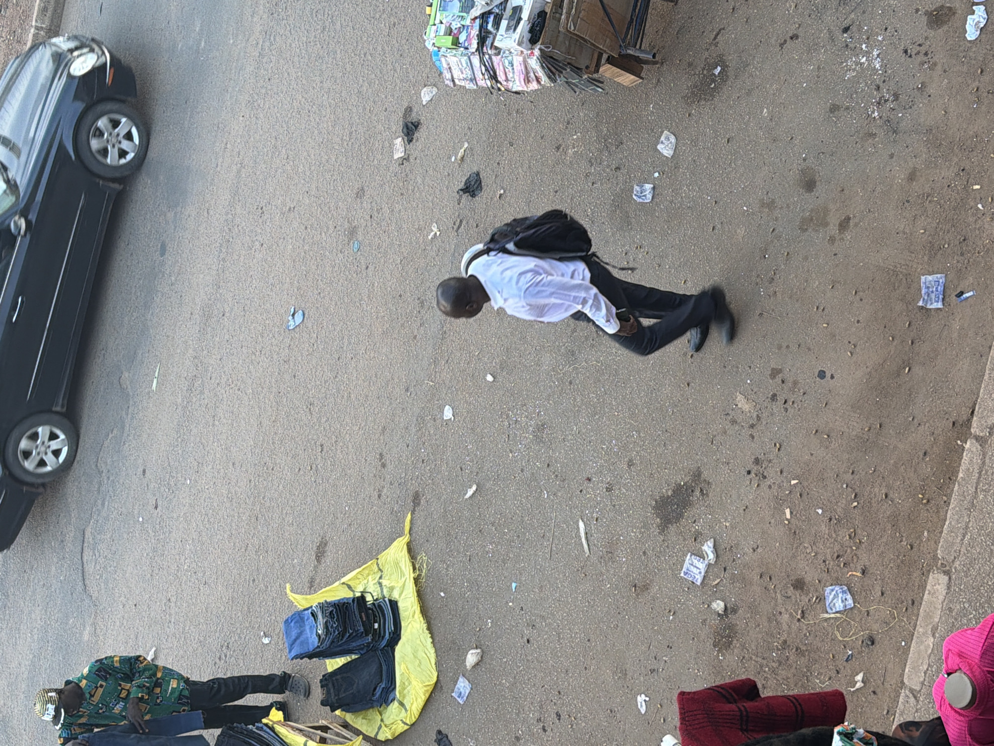 An overhead view captures a street scene in Bukuru, Nigeria, depicting commercial activity and urban elements. A man wearing a green patterned shirt and a striped hat stands near a yellow tarp displaying stacks of folded blue jeans. To his left, a black car with silver rims is parked on the asphalt road. In the middle ground, a man in a white collared shirt and dark pants walks away from the camera, carrying a black backpack. To the right, a wooden cart is laden with numerous folded items, appearing to be textiles or clothing. The ground is a mix of asphalt and dirt, littered with debris including plastic wrappers and other small objects. Clothing items are also visible hanging on the left and right edges of the frame.