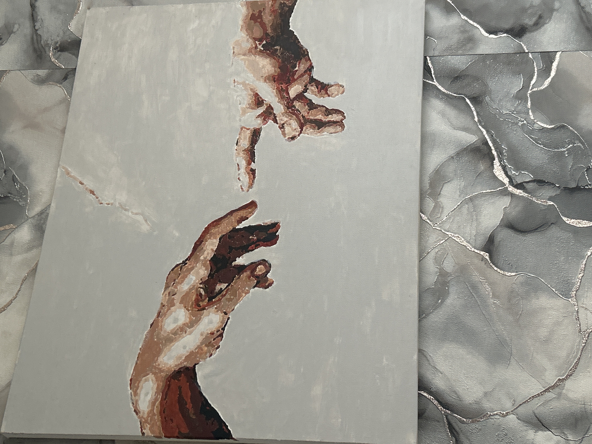 The image shows a painting of two hands reaching towards each other, reminiscent of Michelangelo's "The Creation of Adam." The painting appears to be on a canvas. The background is a neutral gray color. The skin tones are depicted with variations of brown and white paint. The canvas is resting on a surface that has a gray marbled pattern with metallic veining. There is no indication that the location is in Ольховка, Russia.