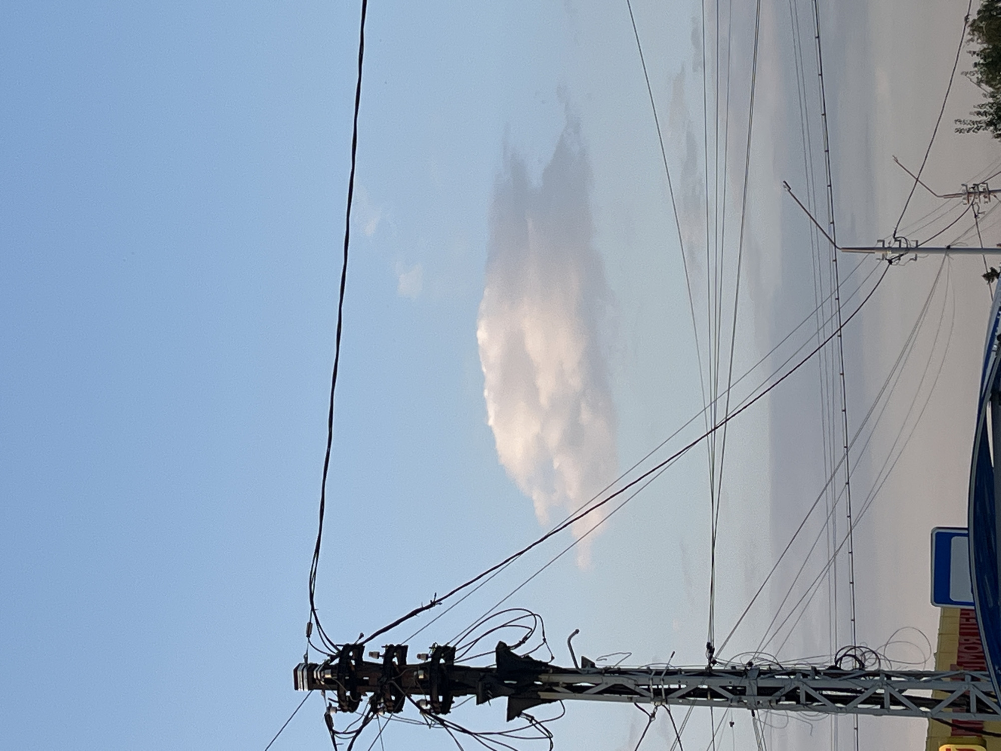 An outdoor, low-angle, medium shot captures a utility pole on the left side of the frame, silhouetted against a pale blue sky with scattered clouds. The pole is adorned with a complex network of wires and cables, suggesting an urban or semi-urban setting. A single, fluffy white cloud is prominently featured in the middle ground of the sky, slightly to the right of the pole. Further in the background, a cityscape is visible, with hints of buildings and trees, characteristic of the city of Saratov, Russia. The lighting suggests it is either early morning or late afternoon.