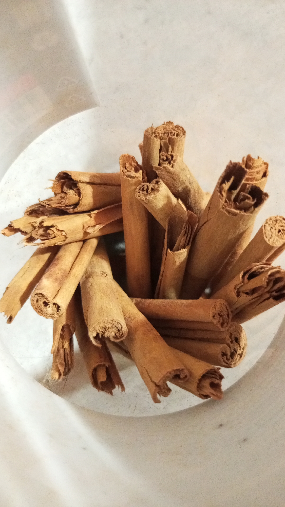 The image shows a close-up, elevated, angled view of a container filled with cinnamon sticks. The sticks vary in size and are tightly packed together, creating a dense cluster. The container appears to be made of a semi-transparent, light-colored material. The background is blurred and indistinct. There are no people visible in the image. The location, Юдино, Russia, cannot be confirmed based on the visual information provided.