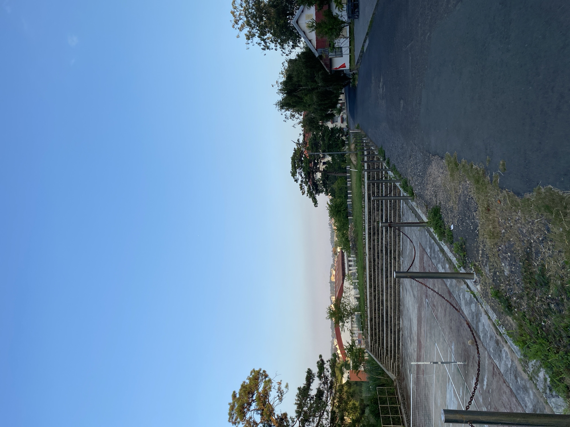 This is a vertical, eye-level, medium shot of a pathway leading uphill in Lâm Viên - Đà Lạt, Vietnam. The pathway is paved with asphalt and lined with metal posts and chains on the left side. To the left of the pathway is a concrete tennis court with a net and a couple of poles. Behind the court are tiered concrete steps. Beyond the steps, there are buildings with red roofs and trees. The sky is a clear, pale blue.
