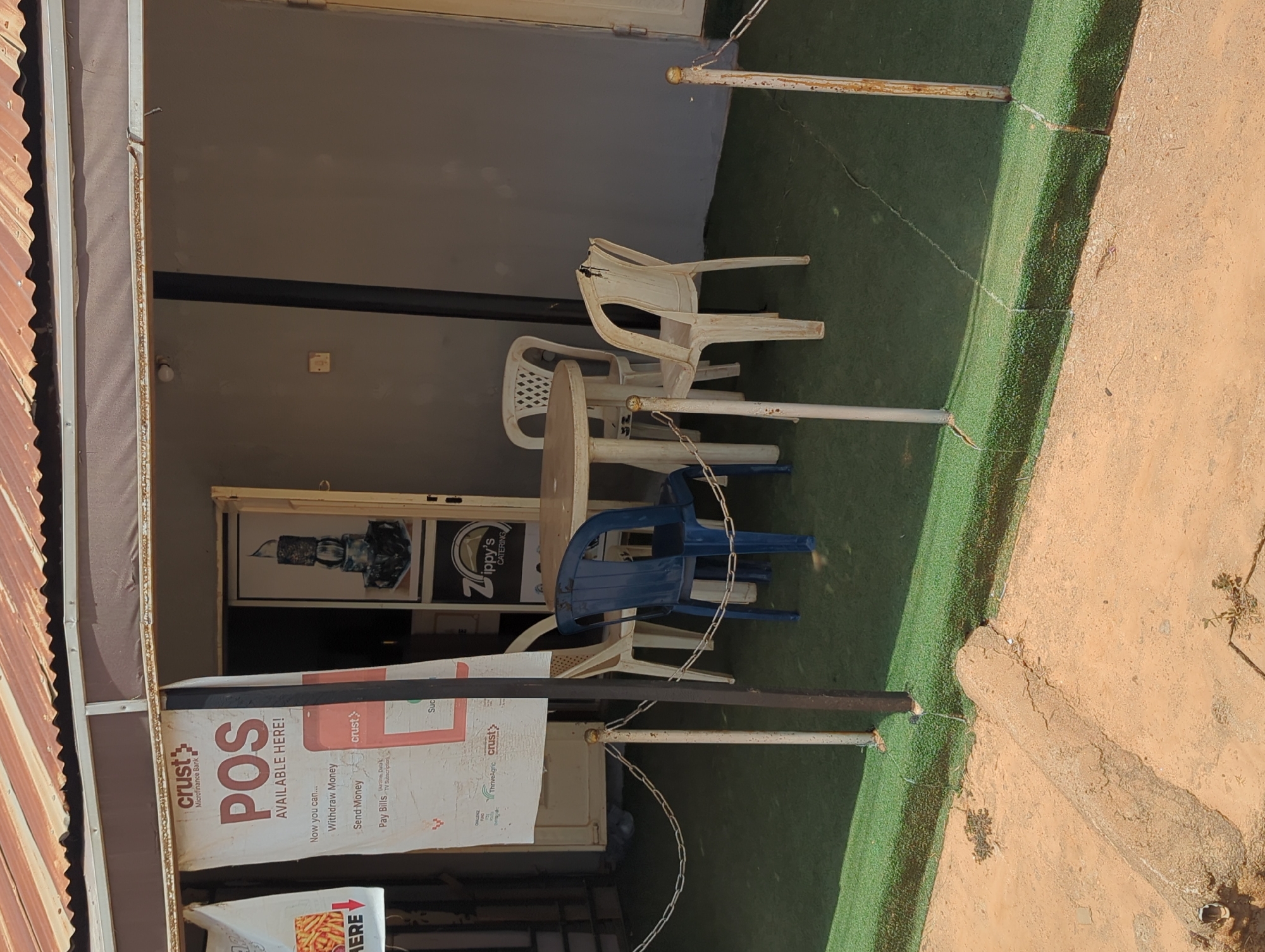 The image depicts an outdoor seating area of a business establishment, possibly a restaurant or cafe, in Narayi, Nigeria. The seating area is defined by a green artificial turf surface. A metal chain fence runs along the edge of the turf, partially obscured by advertising banners. Several plastic chairs of varying colors (white and blue) are arranged around a circular table. A sign indicates that the establishment offers Crust Microfinance Bank POS services, including cash withdrawal, money transfer, and bill payment. In the background, through the entrance of the establishment, a logo for "Zippy's Catering" is visible. The scene is partially covered by a corrugated metal awning.