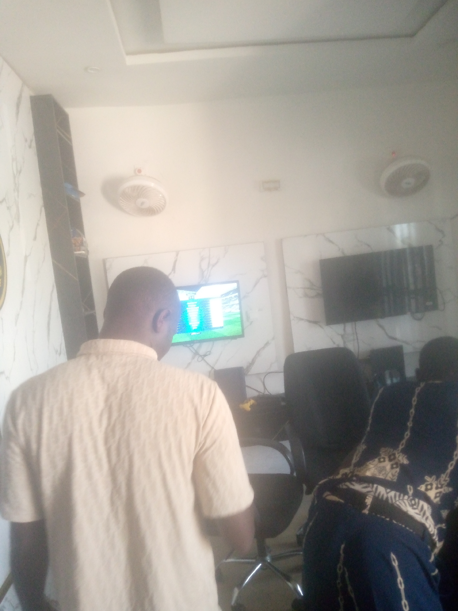 This image depicts an indoor scene in the city of Jalingo, Nigeria, featuring two individuals from behind, facing multiple electronic screens within a contemporary-styled room. The walls are predominantly white, adorned with large rectangular panels featuring a white and grey marble-like pattern. The white ceiling has recessed lighting and distinct ceiling panels. Two white wall-mounted oscillating fans are visible on the upper walls.
On the left wall, a screen displays what appears to be a video game, possibly a football simulation, with a green field and graphical interface elements. To its right, another wall-mounted screen is dark, showing vertical lines, suggesting it is either off or malfunctioning. Further right, partially obscured by the foreground individual, are additional dark monitors on desks.
The person in the foreground on the left is a male with dark skin and short hair, wearing a light-colored (beige or light yellow) short-sleeved collared shirt with a subtle textured pattern. He is positioned facing the screens. To the right, a second individual is partially visible, also seen from behind, wearing a dark blue garment with a light-colored chain-link or patterned design, similarly facing the screens. Black office chairs with metal bases are present around the desks. A tall, dark, open shelving unit is visible on the far left wall, holding a few indistinct items. Cables are visible running behind the desks and monitors.