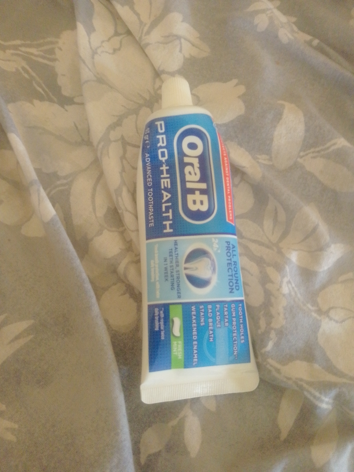 A tube of Oral-B Pro-Health Advanced Toothpaste is positioned on a crumpled, grey and white floral-patterned fabric, likely a bedsheet or duvet cover. The toothpaste tube is mostly blue with white text and features an image of a stylized tooth within a blue circle. The text indicates "ALL ROUND PROTECTION" and lists benefits such as "TOOTH HOLES", "GUM PROTECTION", "TARTAR", "PLAQUE", "BAD BREATH", "STAINS", and "WEAKENED ENAMEL". The tube also states "HEALTHIER, STRONGER TEETH STARTING IN 1 WEEK" and "FRESH MINT". The lighting in the image is soft and diffused. The image does not provide any information about its location in Majemu, Nigeria.
