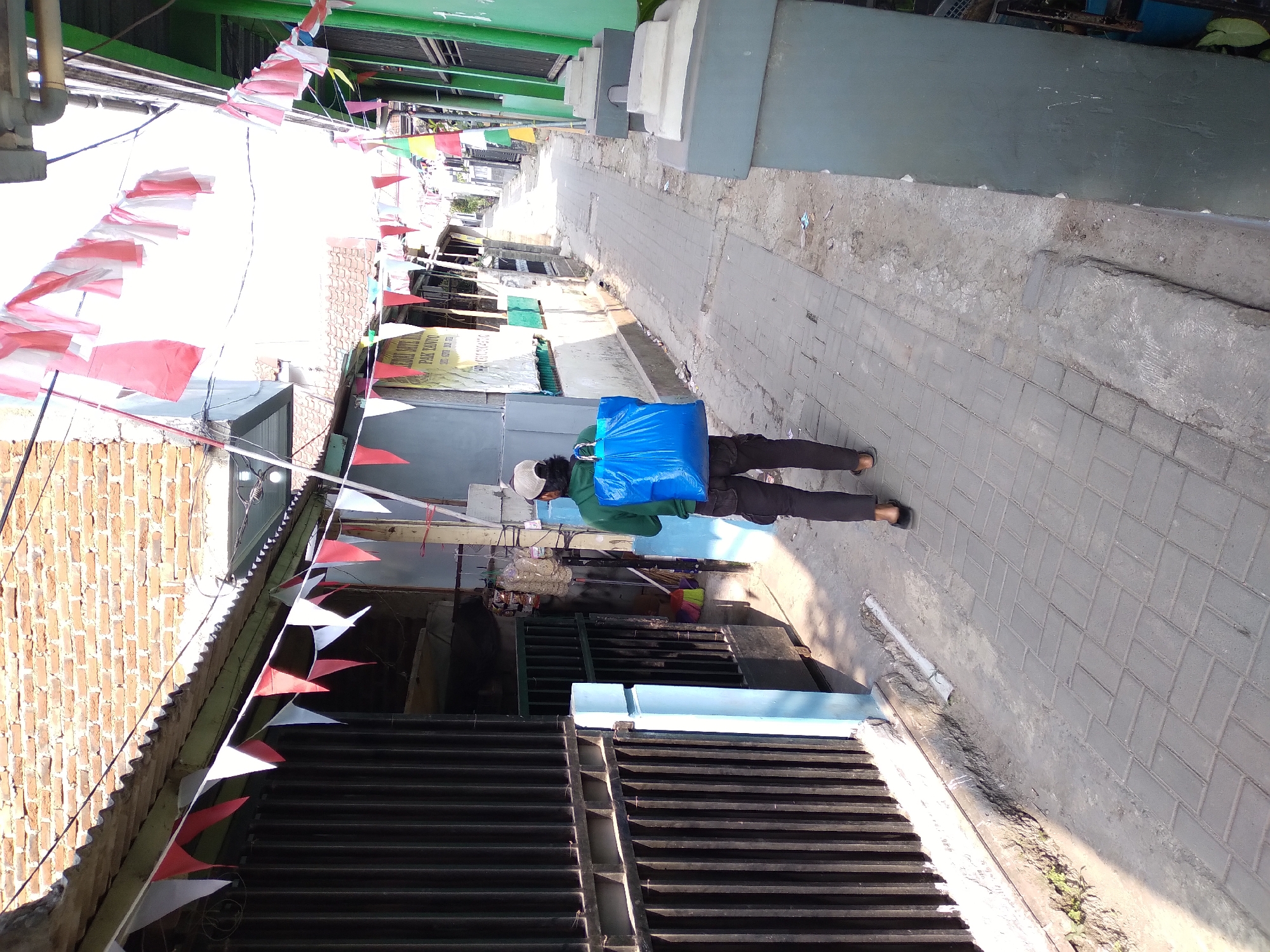 In Cimahi, Indonesia, a person walks down a narrow paved alleyway adorned with red and white triangular flags, likely for a national celebration. The individual, viewed from behind, wears a green long-sleeved shirt, dark pants, and a beige cap, carrying a large bright blue plastic bag over their right shoulder. They are walking away from the camera towards the end of the alley, where more buildings and greenery are visible. The alley is flanked by buildings, one with a corrugated metal roof and brick wall to the left, and a green-painted building to the right. The ground is paved with interlocking concrete bricks, with some areas of dirt and debris. A black metal fence with vertical bars lines the left side of the alley.