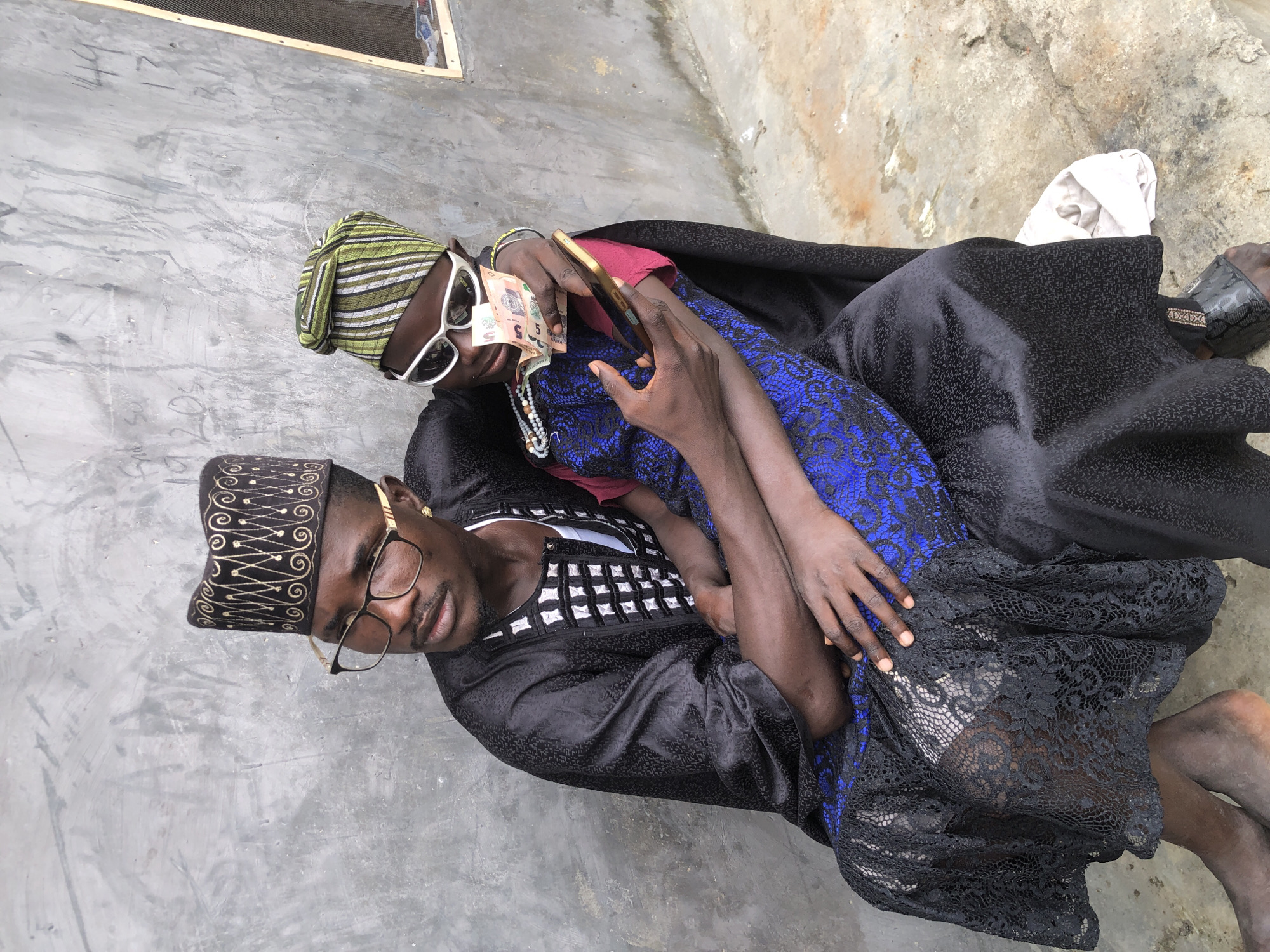 Two young men are sitting in an alleyway in Ado, Nigeria, with a rough, grey concrete wall as the backdrop. The man on the left is wearing a black, ornate cap and a black traditional outfit with white detailing. He has glasses on and is looking off to the side. The man on the right is wearing a green and white striped cap, white sunglasses, and a pink shirt. He is holding several banknotes and a smartphone. The man on the right is sitting in the lap of the man on the left, and the man on the left is holding him. The ground is concrete and appears to be wet in places. There is a small window with a screen to the right of the men.