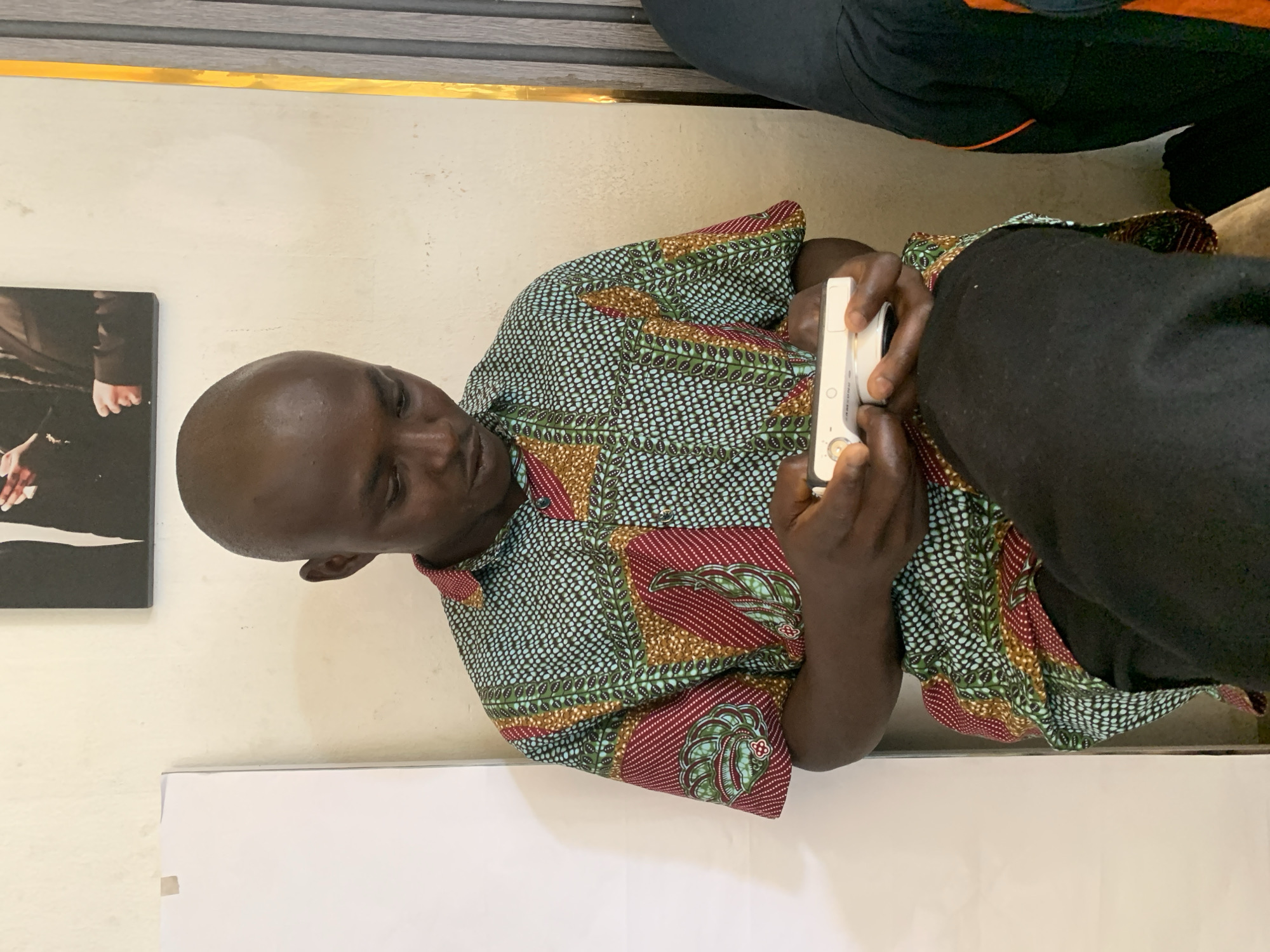 A young Black man with a shaved head is seated in front of a light-colored wall, looking down at a white digital camera in his hands. He is wearing a short-sleeved, button-up shirt with a vibrant, geometric African print featuring shades of green, red, yellow, and blue. His lower body is covered by black pants. To his left, a white board is visible. Above him on the wall, a framed black and white photograph is hanging, showing the upper bodies of two individuals in formal attire. To his right, a vertical golden strip with some light gray vertical paneling can be seen. The lighting in the scene is natural and diffused. The man appears to be engaged in operating the camera. The image seems to be taken indoors, possibly in a room or office setting in Jos, Nigeria, given the context provided.