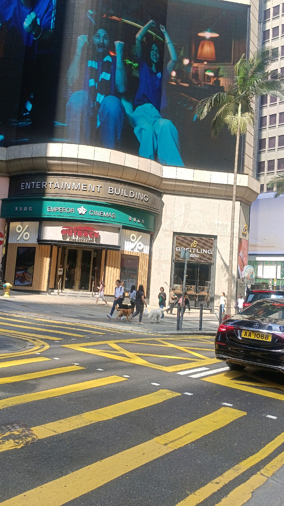 This image shows a street scene in Hong Kong, China, featuring the "ENTERTAINMENT BUILDING" with "EMPEROR CINEMAS" and an "OUTBACK STEAKHOUSE" on the ground floor. A Breitling store is also visible to the right. The street has prominent yellow pedestrian crossing markings and a black Mercedes-Benz car with license plate "AA 1088" is parked on the right side of the road. Several people are walking on the sidewalk, some accompanied by dogs. A large digital billboard is mounted on the building, displaying an advertisement with people. The scene appears to be a busy urban environment with commercial establishments and pedestrian traffic.