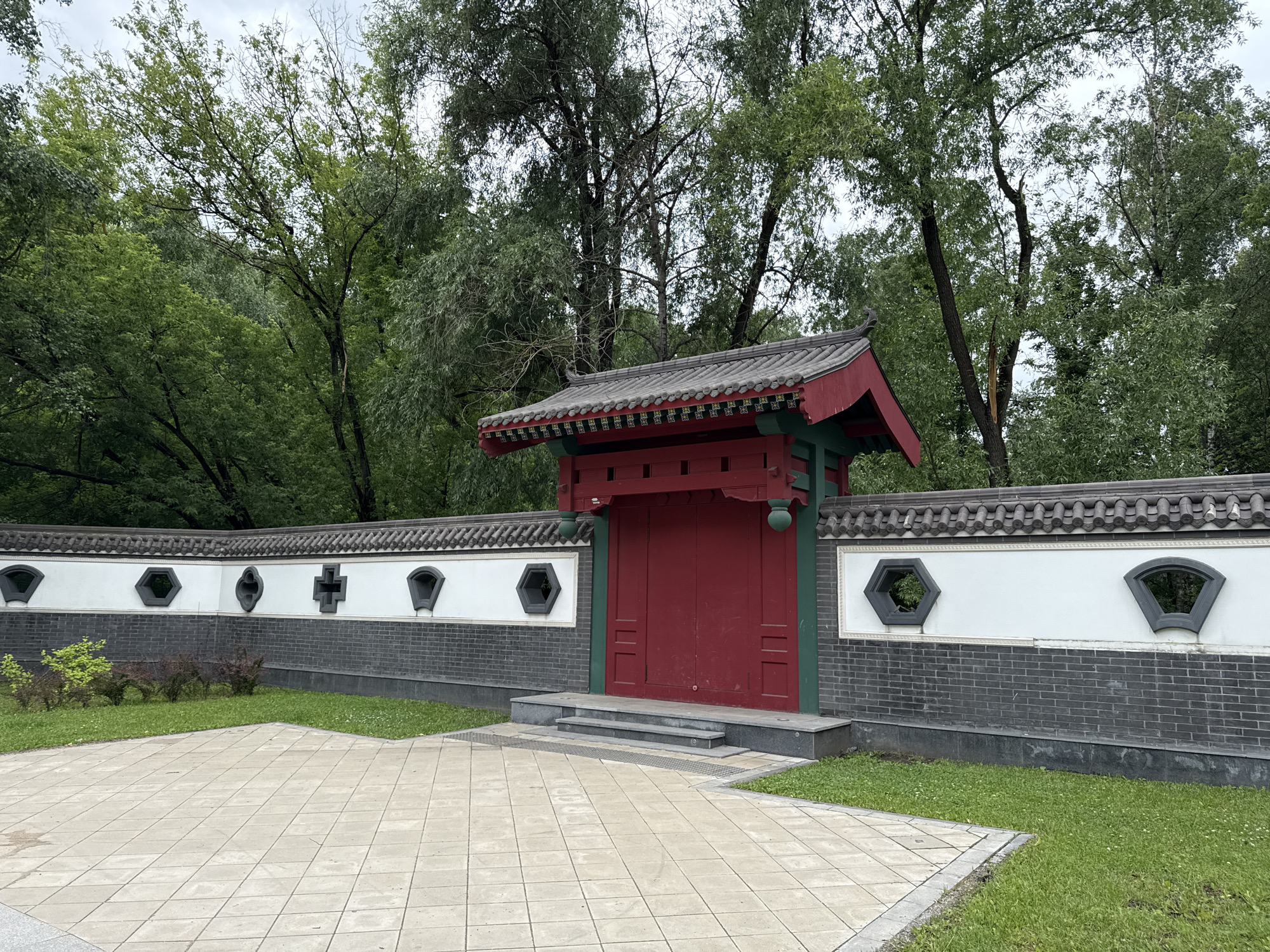 This is a photograph of an ornate red gate set within a traditional Chinese-style wall, located in Moscow, Russia. The gate, painted a vibrant red, is framed by green pillars and topped with a dark, tiled roof with decorative elements. The wall behind the gate is a combination of white plaster and dark brick, adorned with a series of intricately shaped windows. A paved courtyard with a diamond pattern lies in front of the gate, bordered by a strip of green grass. Lush green trees form a backdrop to the entire scene, suggesting a park or garden setting. The sky is overcast, indicating a cloudy day. No people are visible in the image.