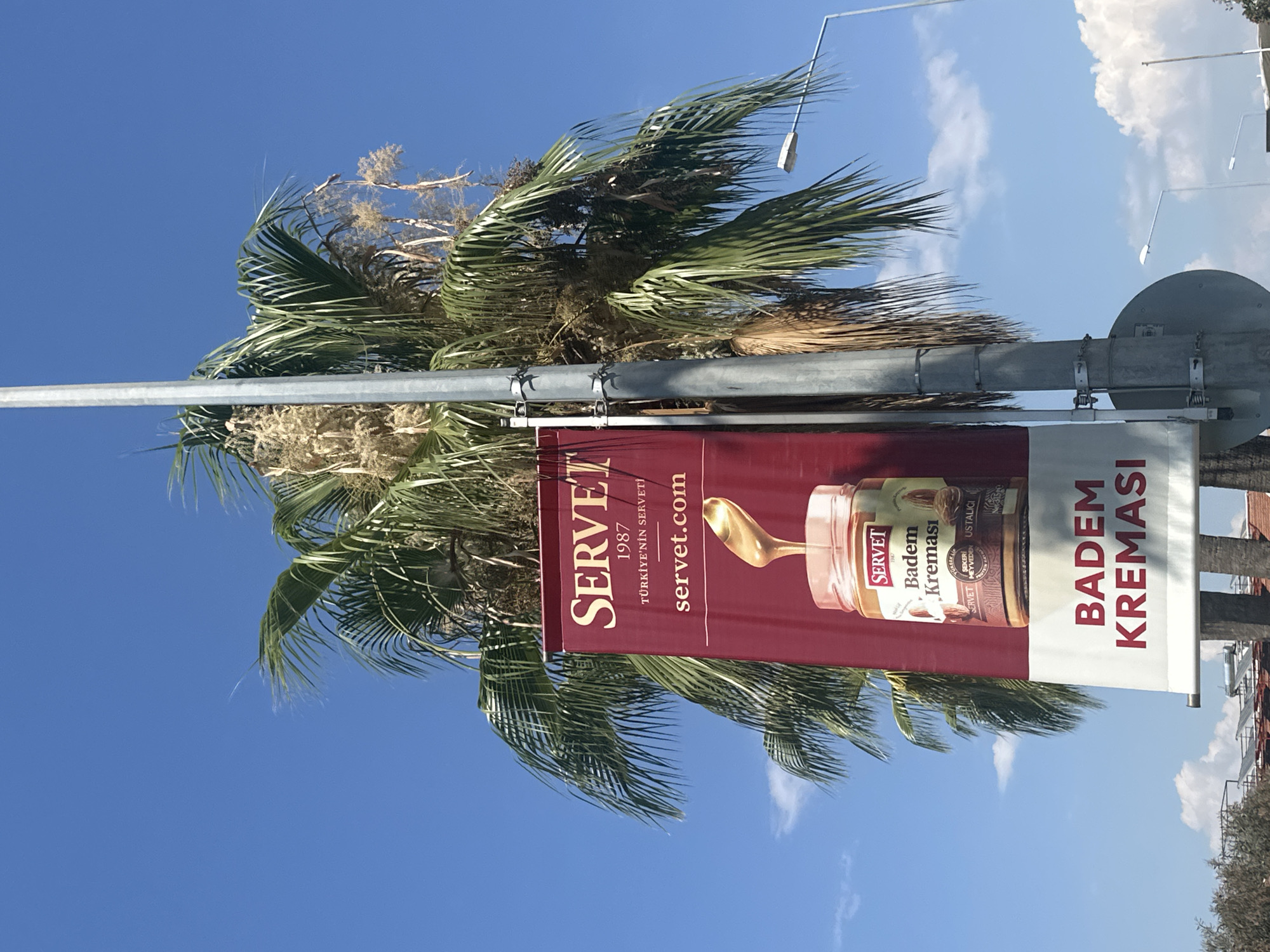 An outdoor banner hangs from a lamppost in Fethiye, Turkey, against a bright blue sky with scattered clouds. The banner, which is predominantly maroon with white lettering, promotes a product called "SERVET Badem Kreması," which translates to "SERVET Almond Cream." The banner displays the SERVET logo, the year 1987, the slogan "TÜRKIYE'NIN SERVETI" (Turkey's SERVET), and the website "servet.com." Below this, an image shows a glass jar of almond cream with a spoon scooping it out. At the bottom of the banner, in large white letters, is the product name "BADEM KREMASI." A large palm tree with green fronds partially obscures the banner. A street light is visible to the right of the lamppost. In the background, some buildings and solar panels are faintly visible.