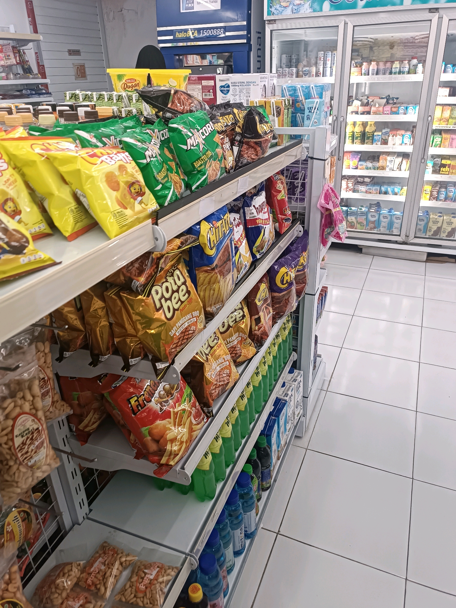 The image depicts the interior of a brightly lit convenience store, likely located in Tabanan, Indonesia, judging by the signage. The foreground is dominated by shelves laden with various snack items, including bags of potato chips and peanuts. Prominent brands like "Pota bee" and "Chitato" are visible. A refrigerated display case filled with drinks occupies the background. The store features white tiled flooring and metal shelving units. The overall impression is of a well-stocked retail environment, typical of a convenience store or minimarket. There are no people visible in the frame.