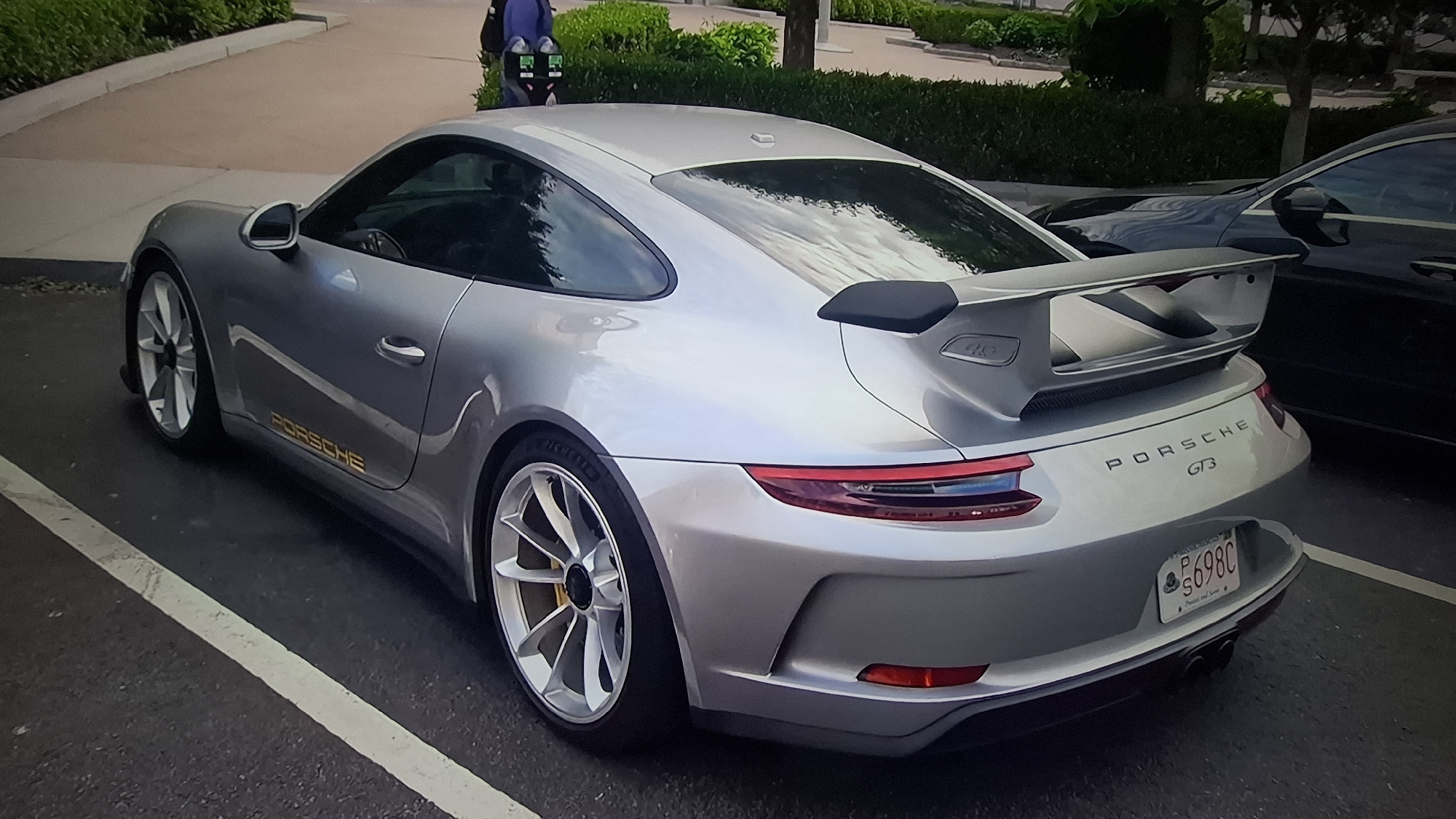 The image displays a silver Porsche 911 GT3, parked outdoors during the daytime. The car is viewed from a rear-three-quarter angle, highlighting its athletic stance and large, fixed rear wing. It features multi-spoke silver wheels with yellow brake calipers visible through the spokes. A gold "PORSCHE" decal is present on the lower side panel of the vehicle. The rear of the car clearly displays "PORSCHE GT3" badging.
A Massachusetts license plate, bearing the alphanumeric combination "S698C", is affixed to the rear. The state name "MASSACHUSETTS" is visible at the top of the plate, and a slogan, "The Spirit of America," can be discerned below.
The Porsche is parked on a paved surface marked with a white line. In the background, a dark-colored car is partially visible to the right. Beyond the parking area, there are green bushes, trees, and a paved walkway. A person, wearing blue and black attire, is faintly visible in the far background, possibly near what appears to be a parking meter or similar fixture. The bright reflections on the car's bodywork suggest clear, sunny, or partly cloudy weather conditions. The scene portrays a high-performance sports car stationary in an outdoor, landscaped parking environment.