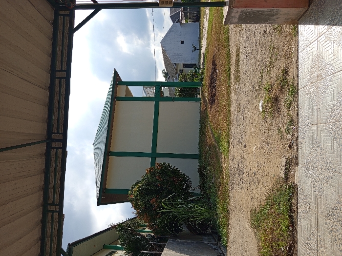 The image depicts an outdoor scene in Keudematangglumpangdua, Indonesia, viewed from beneath a light-colored corrugated metal canopy supported by dark metal framework. In the immediate foreground, a patterned tiled surface is visible, extending to the bottom of the frame. A concrete pillar is partially visible on the right. The midground consists of an unpaved area of earth, sparsely covered with green grass and patches of dry, reddish-brown vegetation.
In the background, a two-story building with cream-colored walls and prominent green structural elements stands on the left. It features a dark green corrugated hip roof. A large, rounded green shrub with some reddish foliage is situated in front of this building, accompanied by taller green plants. Further to the right, a smaller, single-story white building with a dark roof is visible. The sky is overcast with a mix of darker clouds and brighter areas. No individuals are present in the scene.