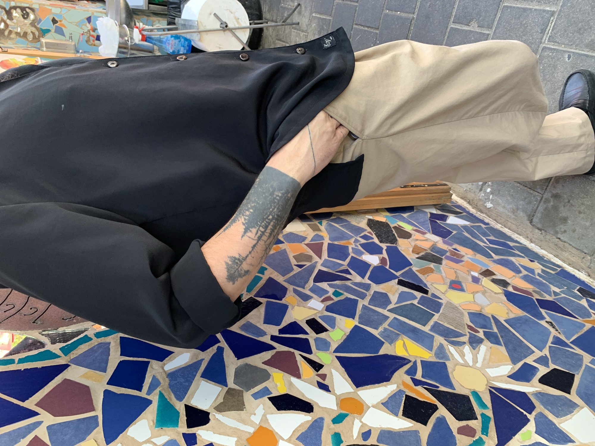 A man is seated on a mosaic-tiled surface, likely a bench or table. He is wearing a black button-down shirt and tan pants. His left arm, which is visible, has two tattoos: one consisting of a dark band around the wrist and another depicting a forest scene on his forearm. He is sitting with his legs to the right of the frame and his hand partially inside the right pocket. The mosaic tiles are predominantly blue with accents of yellow, orange, white, and other colors, forming an abstract pattern. In the background, there are hints of a street scene, including a water bottle and other indiscernible objects. The location of the image is in the city of Tel-Aviv, Israel.