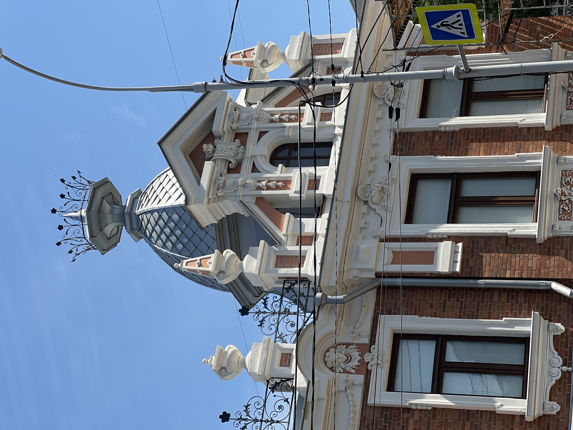 The image shows a close-up, low-angle shot of the ornate upper facade of a brick building in Krasnodar, Russia, under a clear blue sky. The architecture is characterized by a domed roof with intricate metalwork, decorative moldings, and sculpted embellishments above arched windows. A prominent street lamp stands to the right, adorned with numerous black electrical wires. A pedestrian crossing sign, a blue square with a white pedestrian symbol, is attached to the lamp post. The building's brickwork is a warm reddish-brown, contrasting with the white and pale pink decorative elements. The overall impression is one of historical architecture with a touch of grandeur.