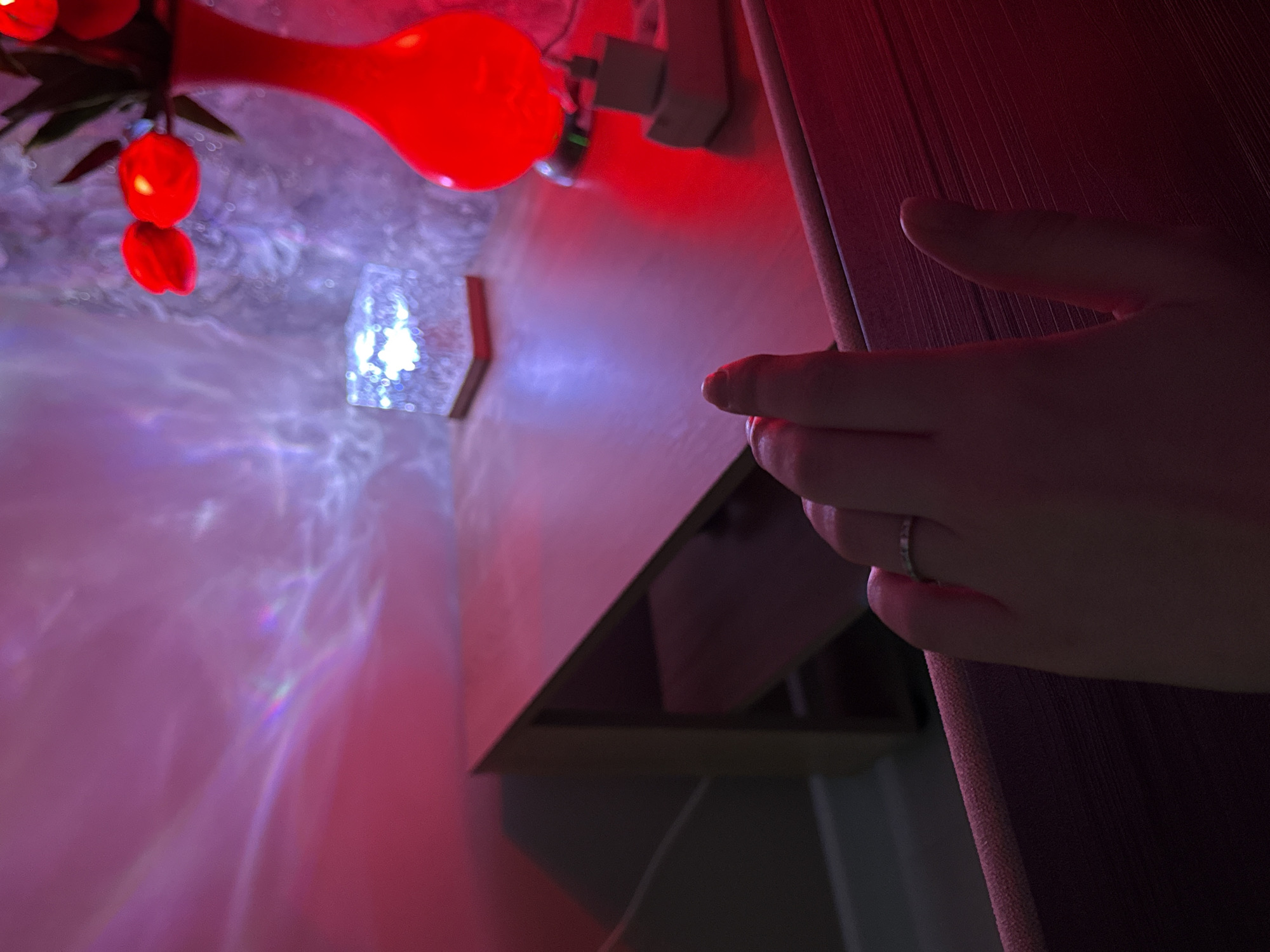 A close-up indoor shot shows a human hand wearing a ring partially obscuring a cabinet door. A red artificial flower arrangement and a small, glowing, crystalline decoration are visible on the left, casting refracted light patterns on the surrounding surfaces. The overall lighting is a mix of red, purple, and white. The setting appears to be in a domestic environment, but the precise location is not discernible from the image.