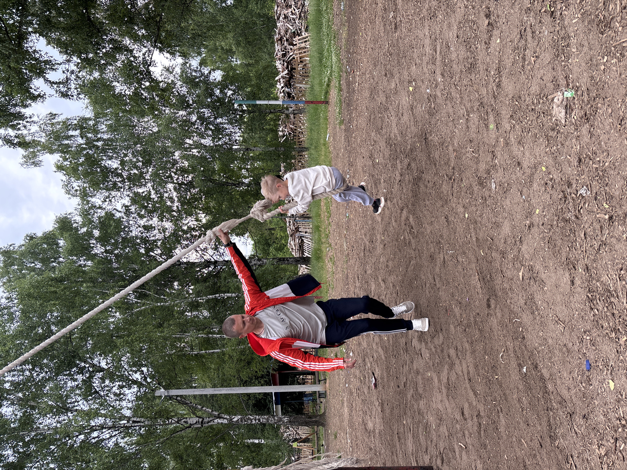 A man in a red and white striped jacket is holding a rope swing for a young boy who is swinging. The scene appears to be in a park or outdoor recreational area in Chaikovsky, Russia. The ground is covered in dirt and wood chips. Trees and a cloudy sky are visible in the background.