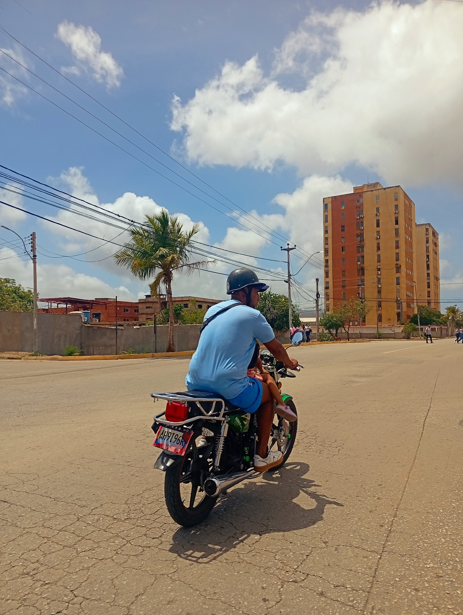 A man wearing a blue t-shirt and blue shorts, with a black helmet, rides a motorcycle down a cracked asphalt street. He has a young child seated behind him, wearing pink shoes. The motorcycle has a license plate reading "AH9796K". In the background, two tall apartment buildings, one orange and one beige, stand under a blue sky with scattered white clouds. Power lines stretch across the sky. To the left, there is a palm tree and a low concrete wall with a building under construction behind it. The scene appears to be in an urban environment.
