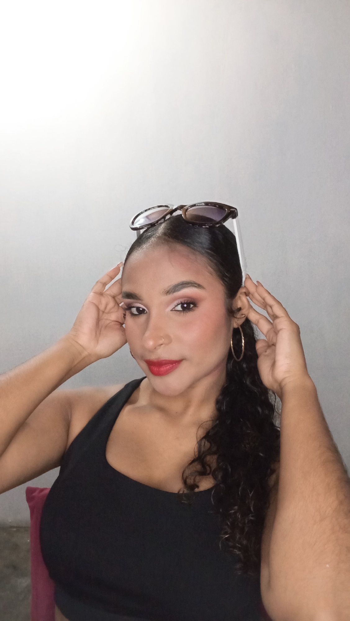 A woman poses indoors, possibly in a residence. She is wearing a black tank top and gold hoop earrings, with her hair styled up with sunglasses perched on top of her head. Her hands are raised near her face, framing it. She wears makeup.
The environment has a plain, light-colored wall. The location cannot be determined without additional data.