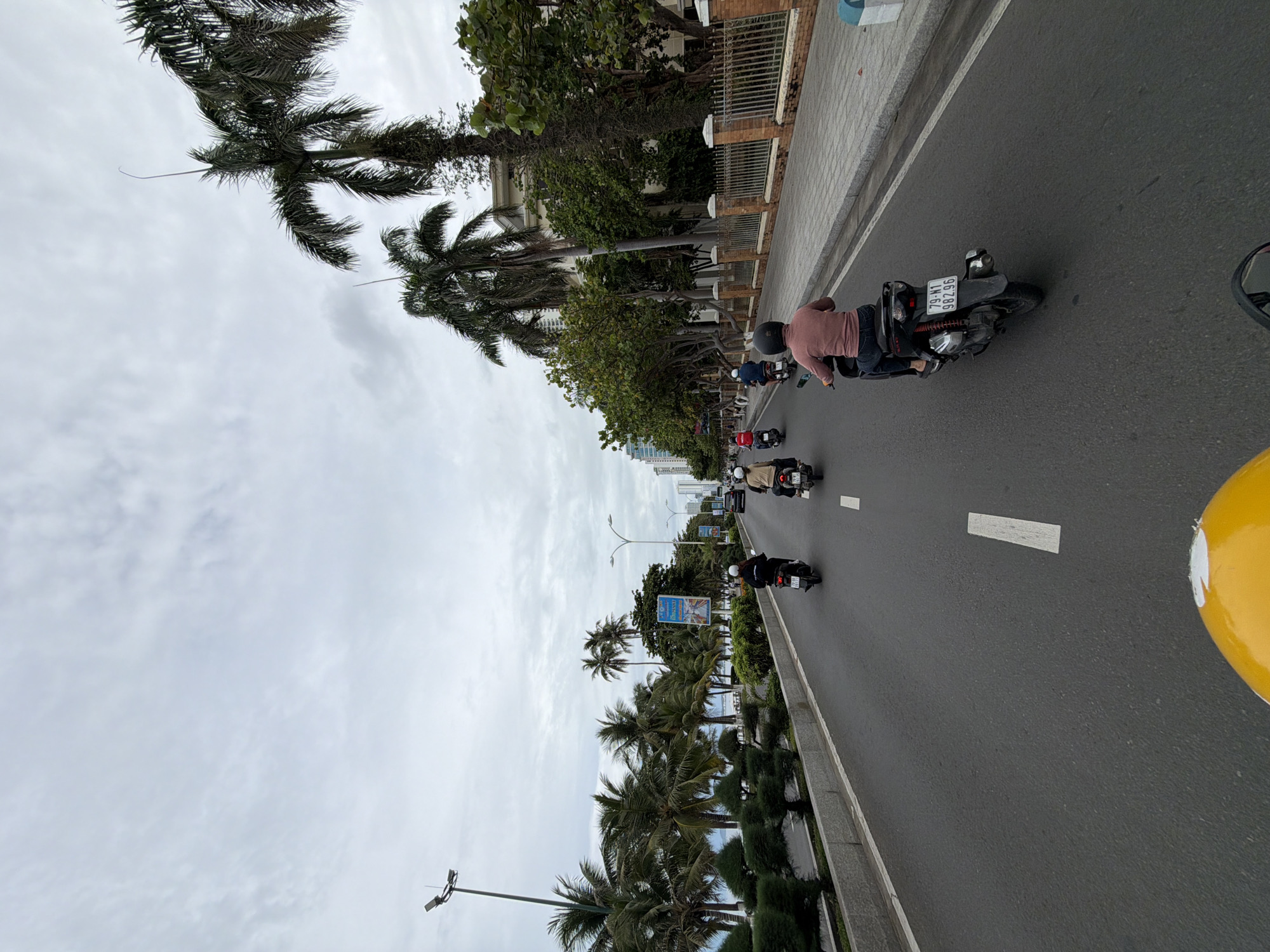 The image depicts a street scene in Nha Trang Ward, Vietnam, viewed from a high angle. Several individuals on motorbikes are traveling along a multi-lane road. Palm trees line one side of the road, while buildings and other vegetation are visible on the opposite side. The sky is overcast. A large yellow object is visible in the lower right corner of the frame. The motorbike closest to the camera bears license plate 79-N1 98296. A few road markings are present on the asphalt surface.