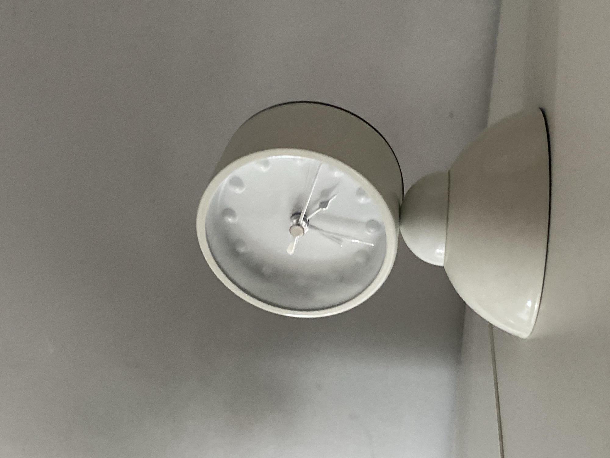 A white, modern-style alarm clock with a rounded base and a tilting clock face sits on a white surface against a plain, light grey wall. The clock face is circular with small raised dots marking the hours. The clock hands are thin and metallic. A thin white cord is visible extending from the base of the clock towards the left side of the frame. The lighting in the scene is soft, creating a subtle shadow on the wall behind the clock. There are no people present. The image is taken from a slightly low angle. This scene is not in Wrocław, Poland.